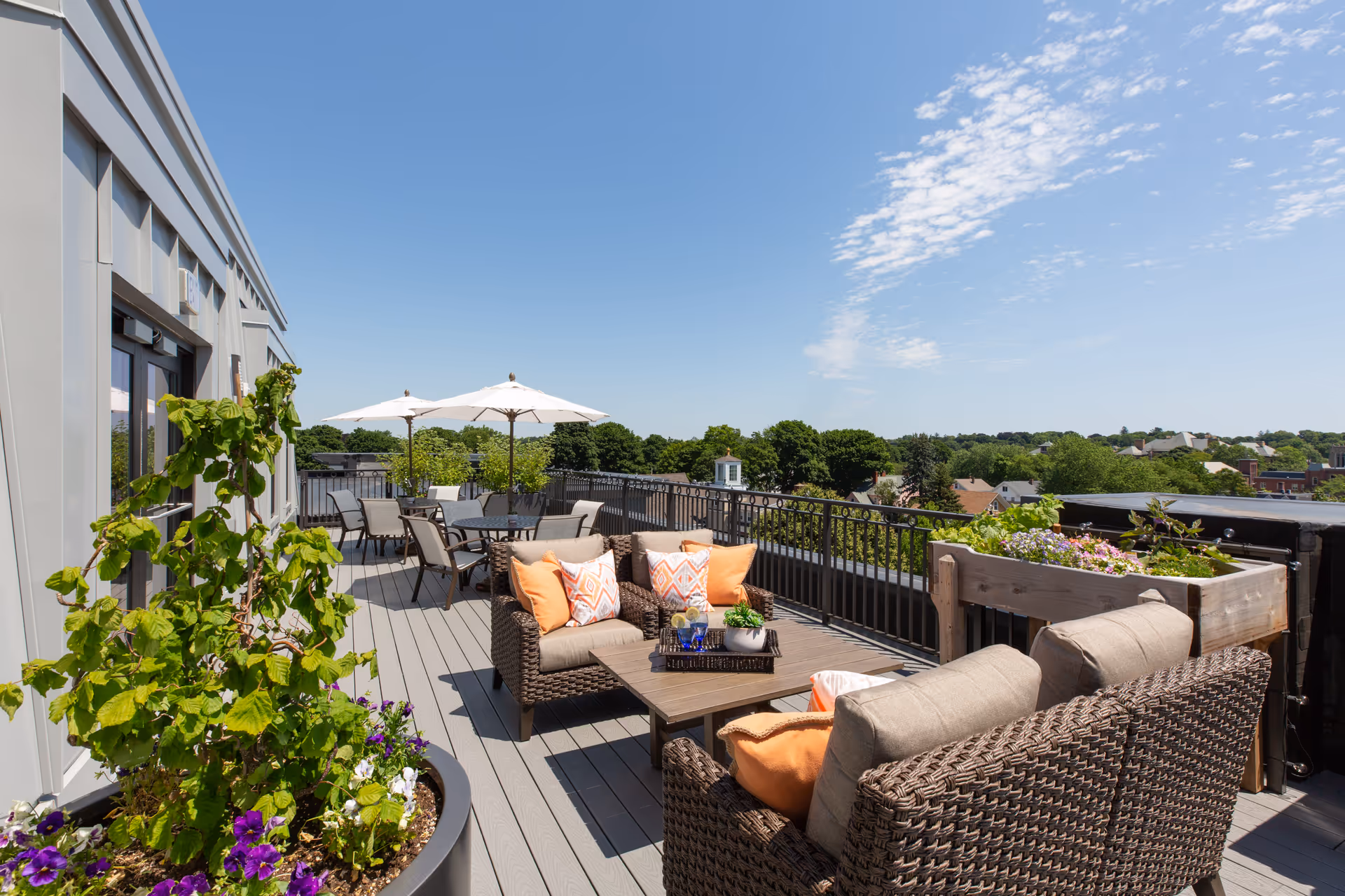 Rooftop terrace with wicker seating, dining tables under umbrellas, potted plants, and a view over treetops.