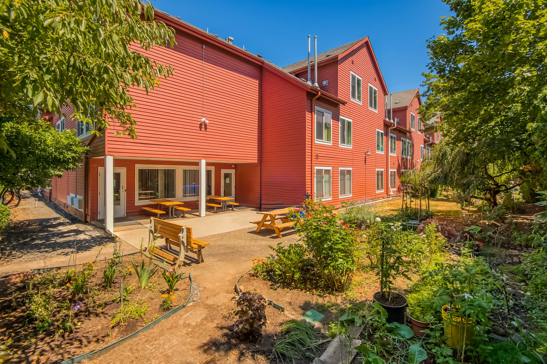 Red three-story building with a patio of picnic tables and a planted garden in the foreground under a clear blue sky.