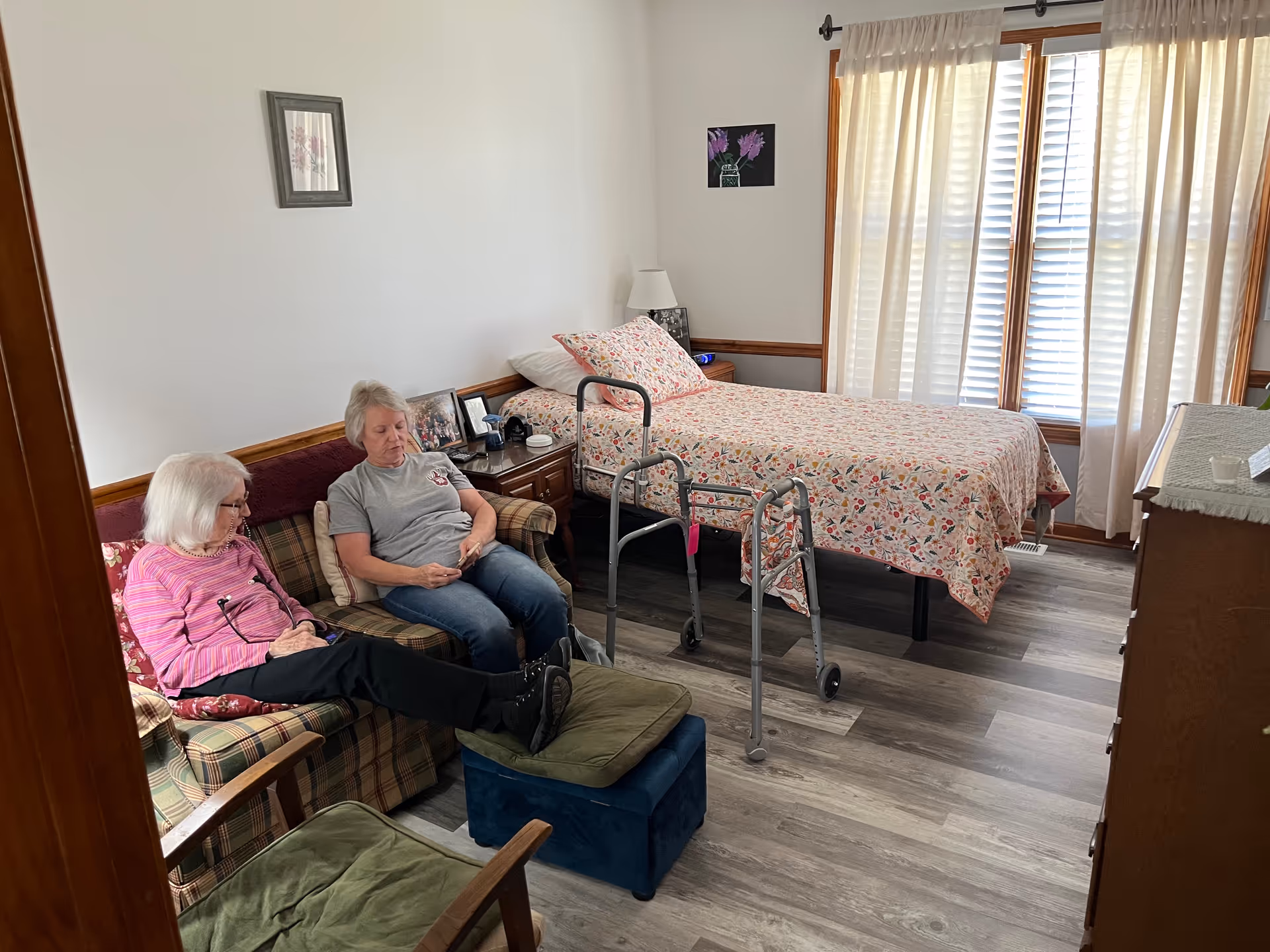 Two elderly residents sit on a sofa in a bedroom suite with a bed and walker.