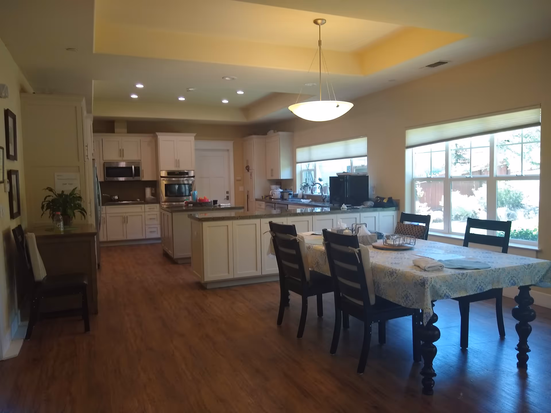 Open-concept kitchen and dining area in a senior living facility with white cabinets and large windows.