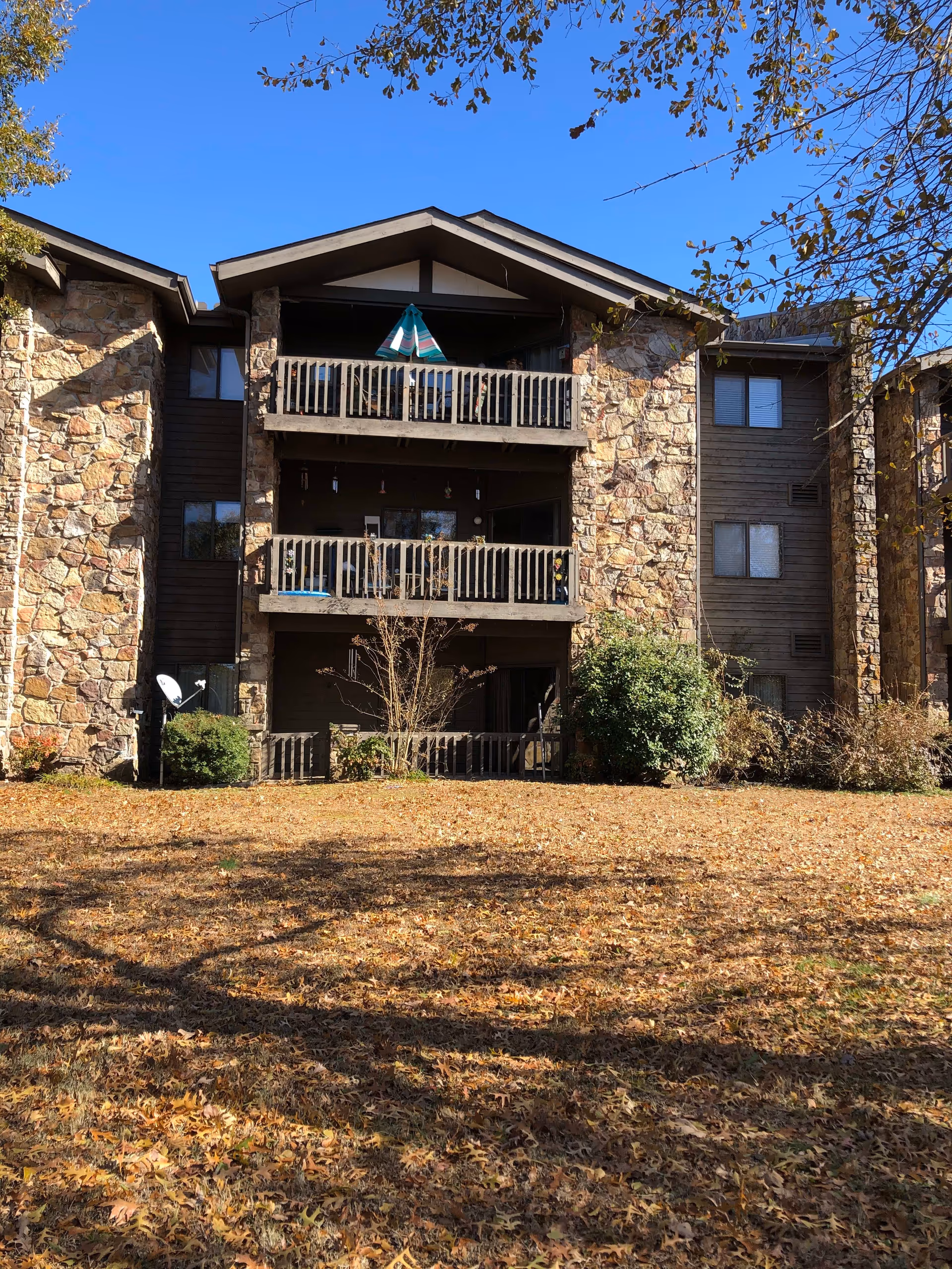 Exterior view of a three-story residential building with stone and wood siding. The building features balconies on each floor with railings, some decorated with plants and a colorful umbrella. The foreground shows a lawn covered with fallen autumn leaves, and trees with sparse leaves frame the image under a clear blue sky.