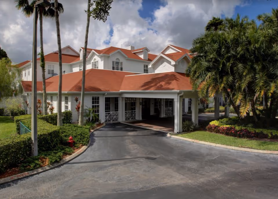 Front entrance of a multi-story white building with red roofs, a covered porte-cochère, palm trees and landscaped driveway.