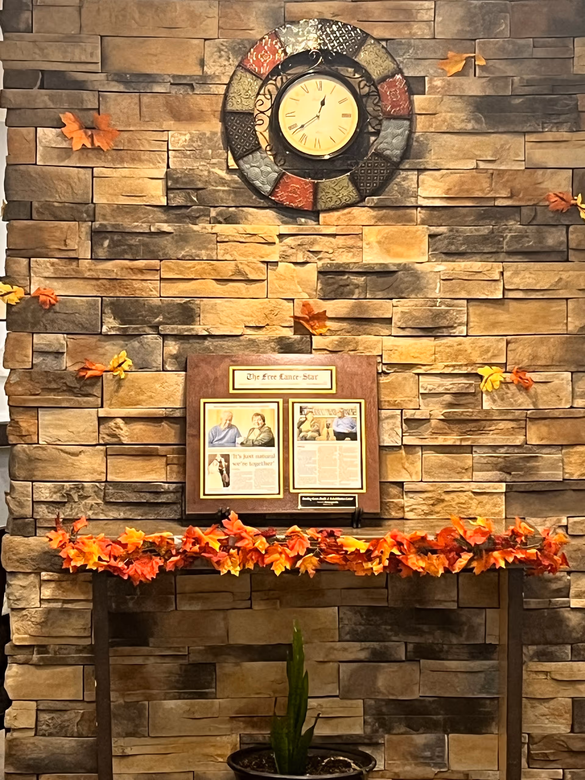 Stone wall interior with round clock, autumn leaves garland, and framed display on table.