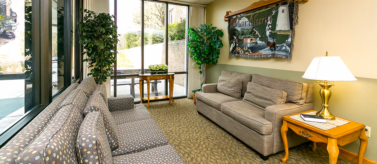 A cozy sitting area in a nursing and rehabilitation facility featuring two sofas with patterned and plain cushions, a wooden side table with a brass lamp and a book, two large potted plants, a small wooden table near large windows with vertical blinds, and a decorative wall hanging depicting Maryland.