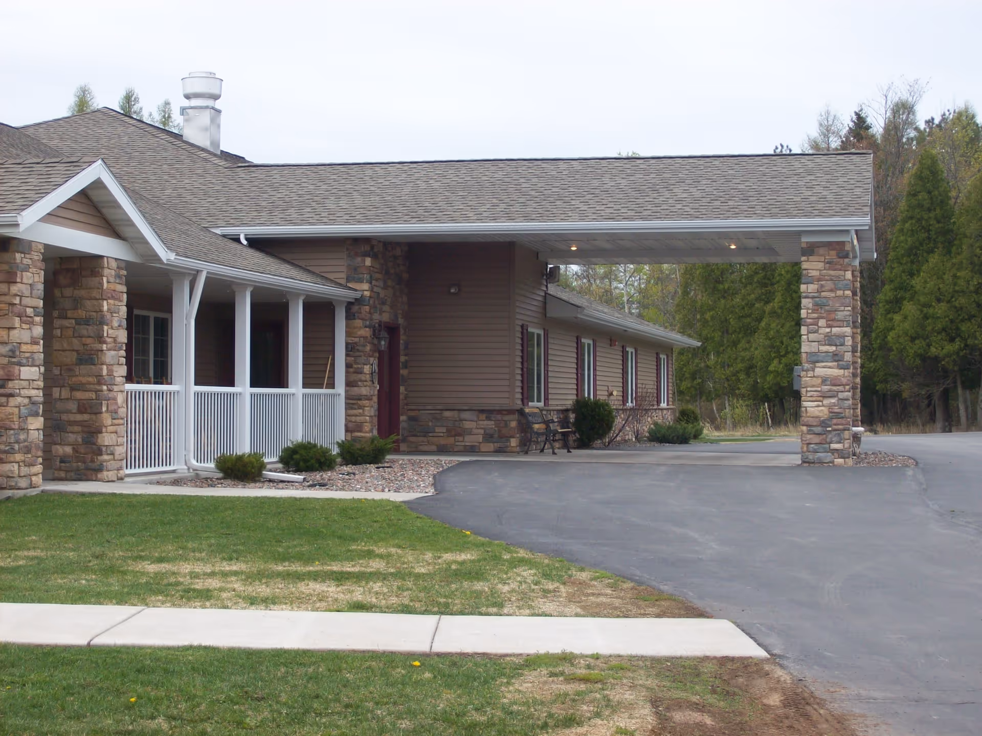 Front entrance of a single-story senior living building with a covered porte-cochère, stone columns, a porch and driveway.