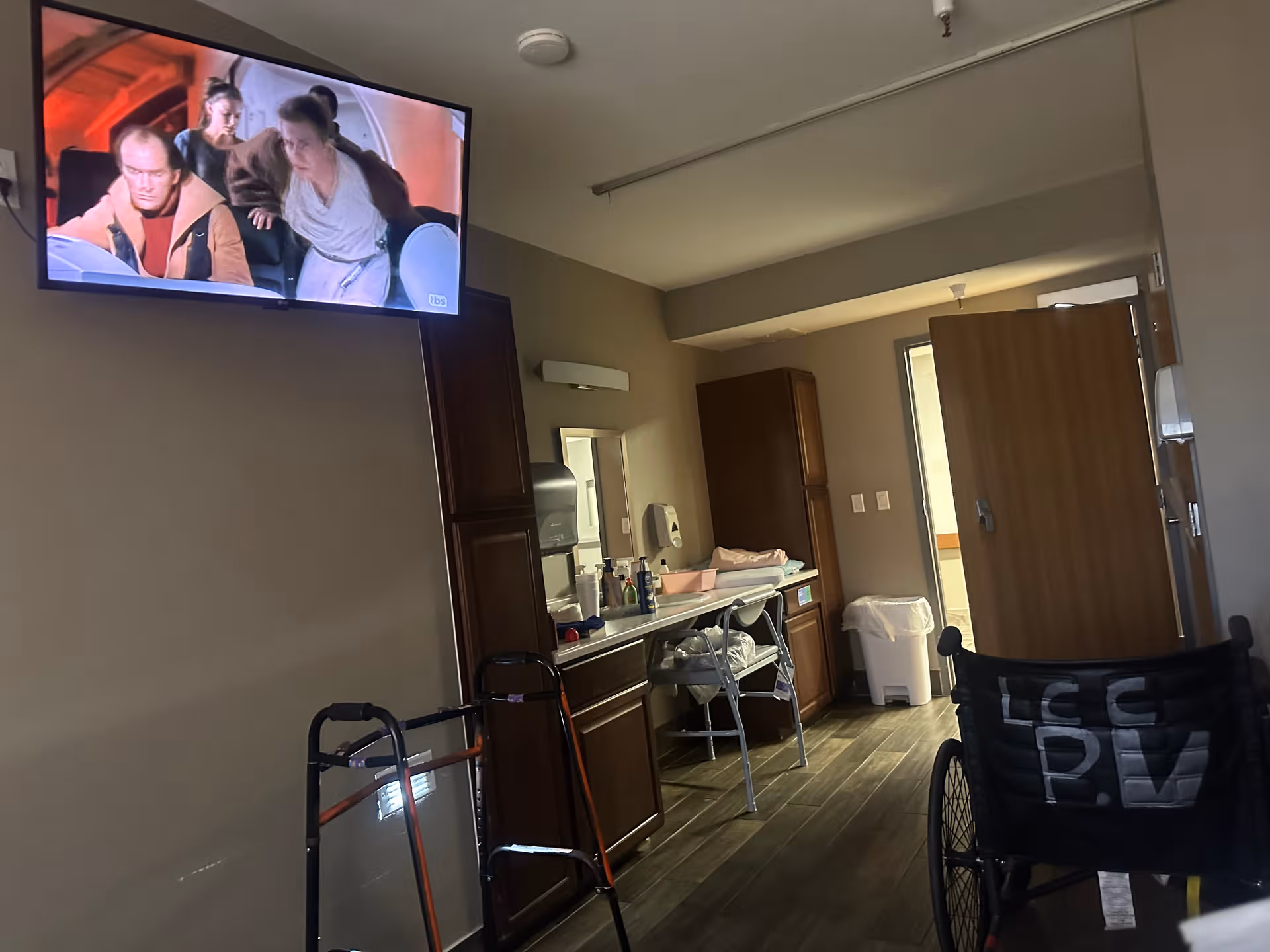 Interior living area with mounted TV, dark cabinets, walker, and wheelchair in view.