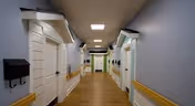 A clean, well-lit hallway in a senior living facility with light blue walls, wooden flooring, white doors on both sides, and yellow handrails along the walls.