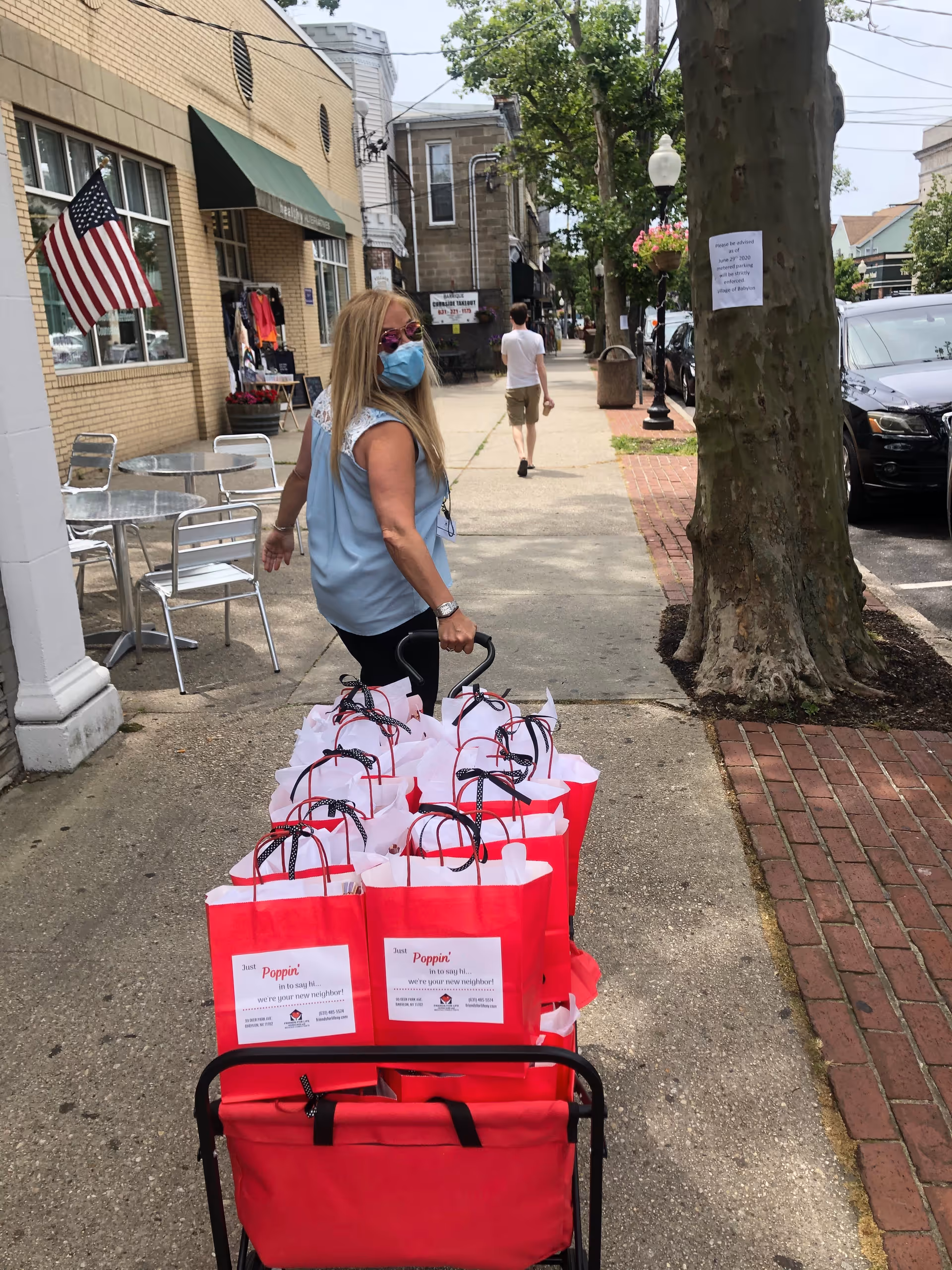Masked woman pushes a cart filled with red gift bags along a sidewalk outside storefronts.
