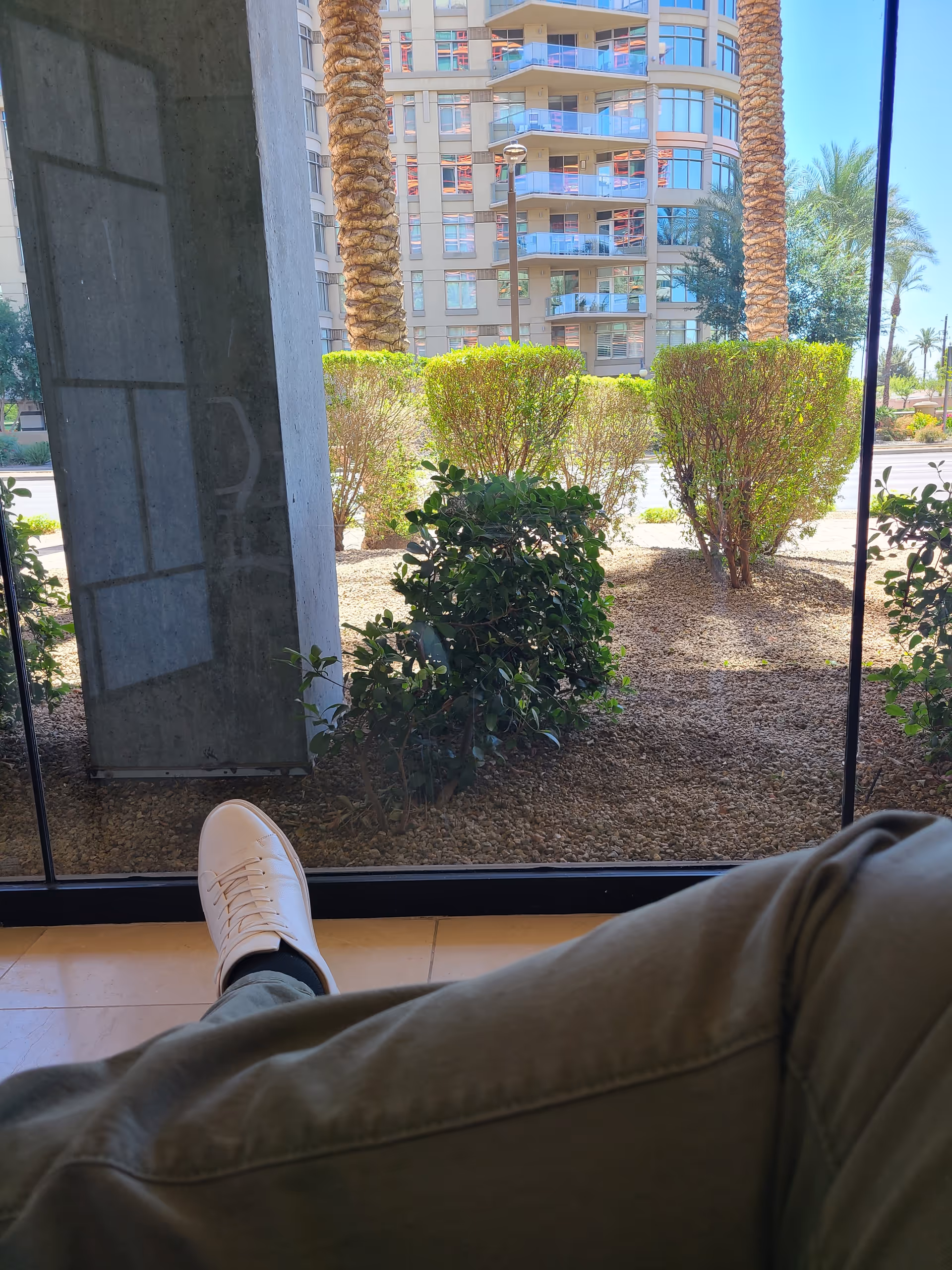 Inside a building, a person’s white sneaker rests by a glass wall overlooking palm trees.