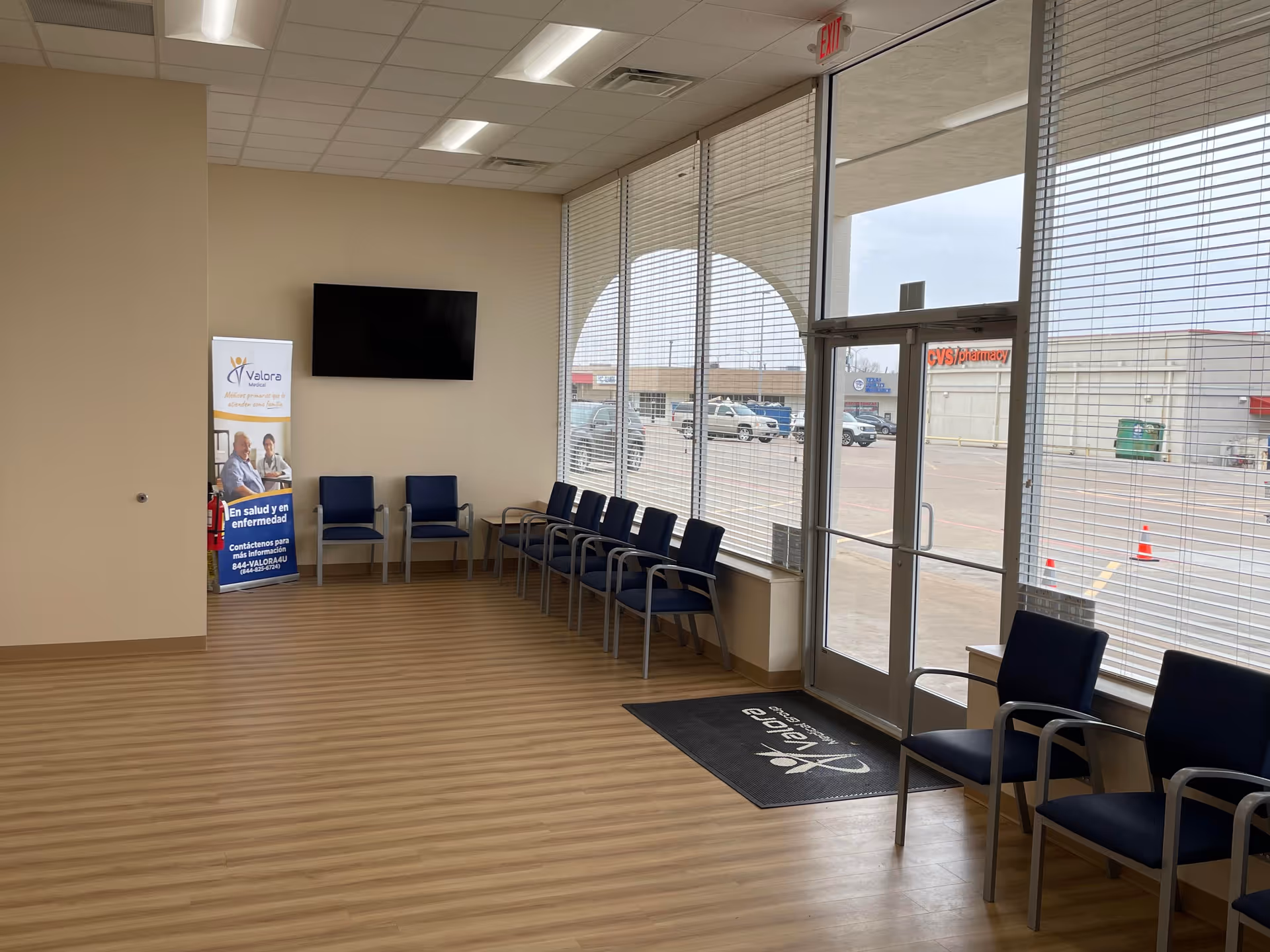 Indoor waiting area with blue chairs, a wall-mounted TV, a banner, and large windows.