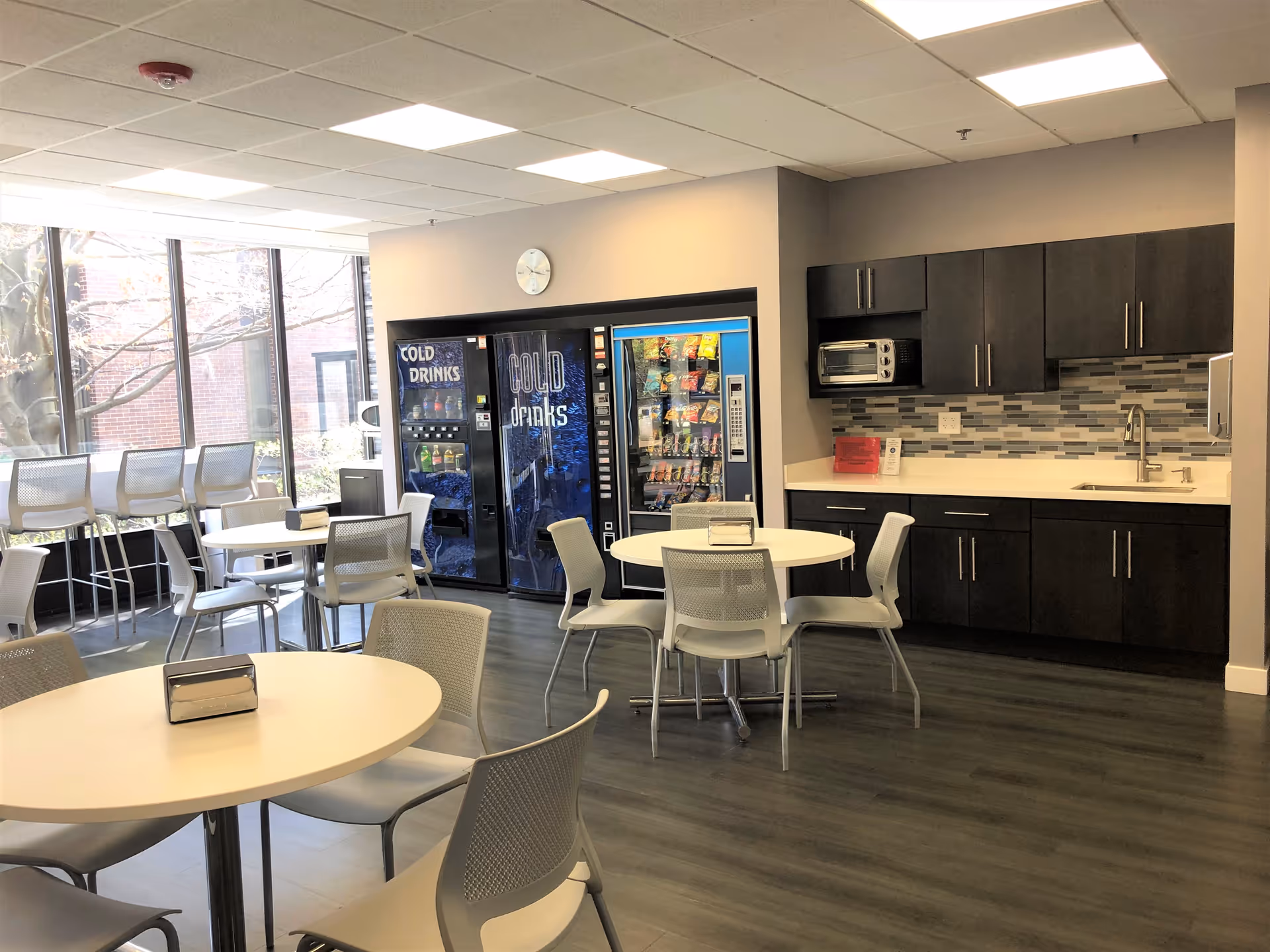 Bright shared dining area with round tables, chairs, vending machines, and a compact kitchen.