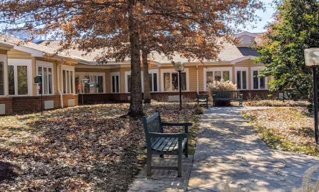 Outdoor courtyard with a paved walkway, benches, trees and fallen leaves in front of a single-story building with many windows.
