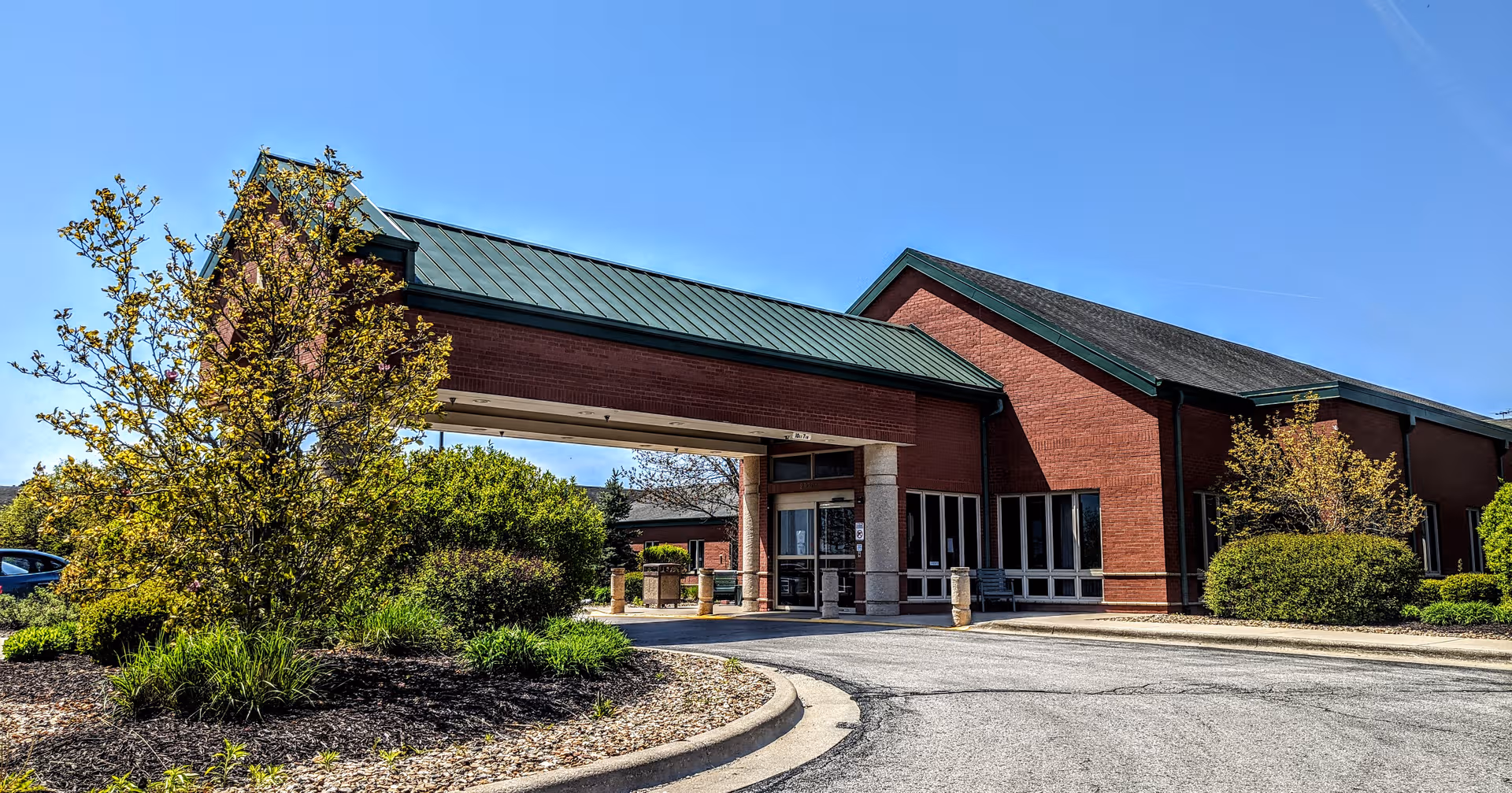Brick senior living center with green roof, covered entrance, and landscaped surroundings.