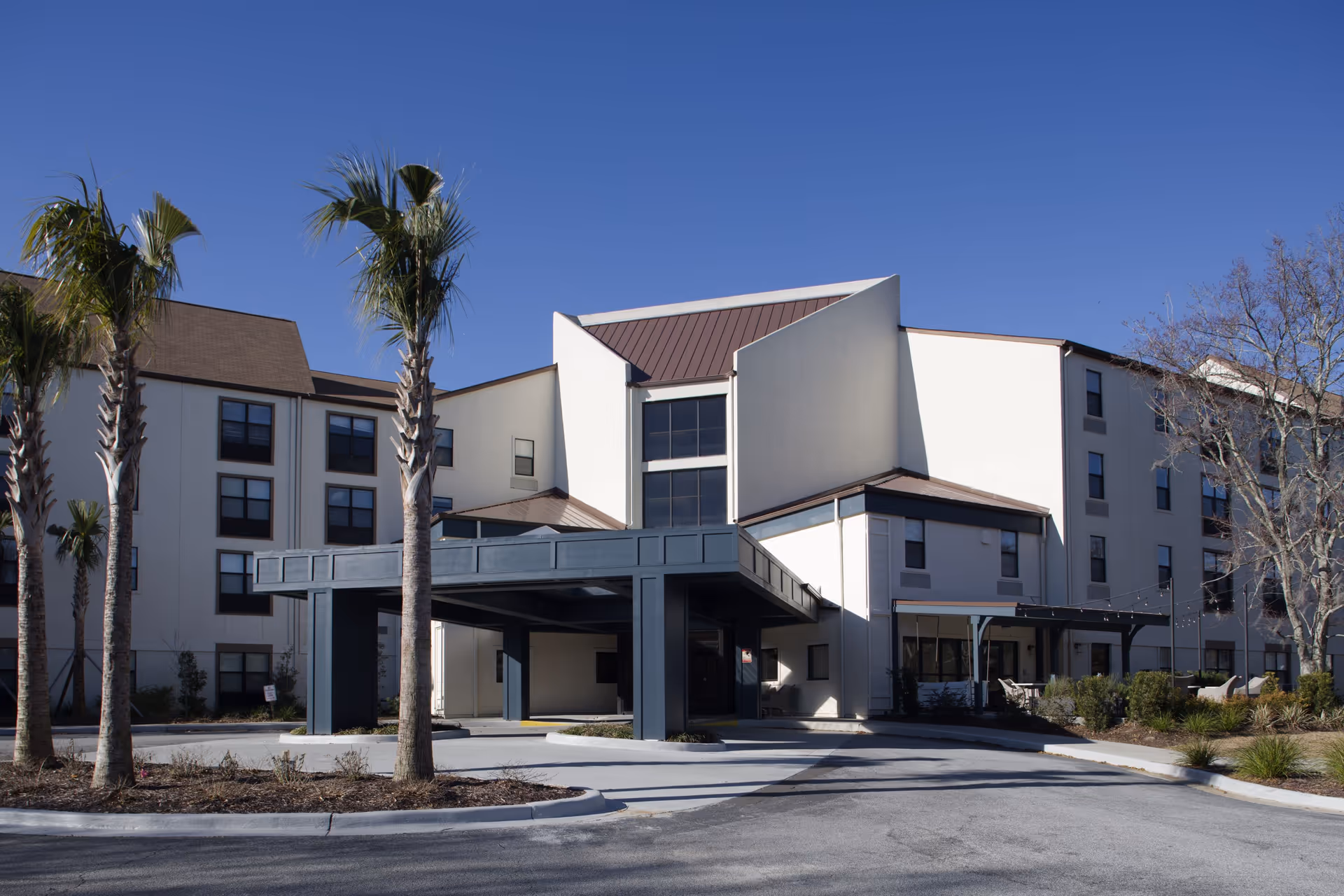 Exterior view of a multi-story senior living facility building with a covered entrance, palm trees, and clear blue sky.