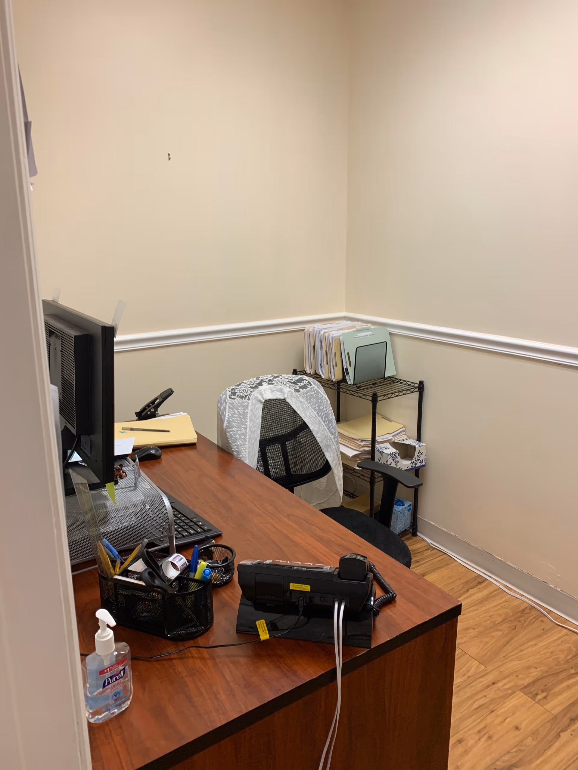 A small office desk with computer, files, and hand sanitizer in a beige interior.