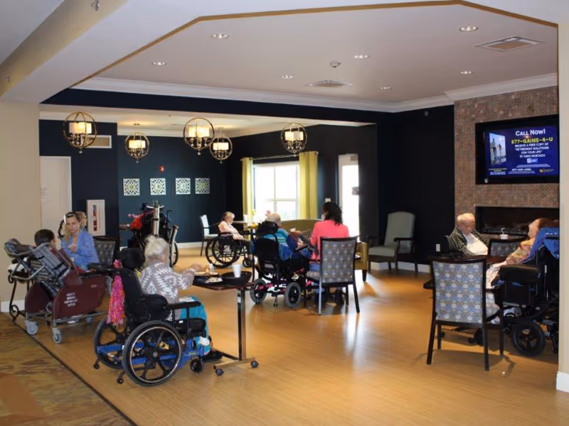 Senior living common room with residents in wheelchairs seated at tables and a television on the wall.