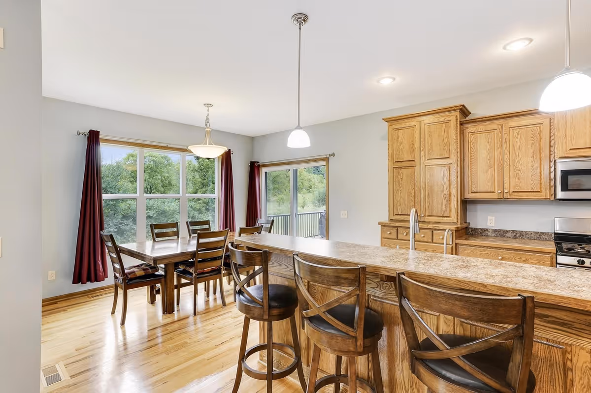 Open-concept kitchen with wooden cabinets, island, bar stools, and a dining area by windows.