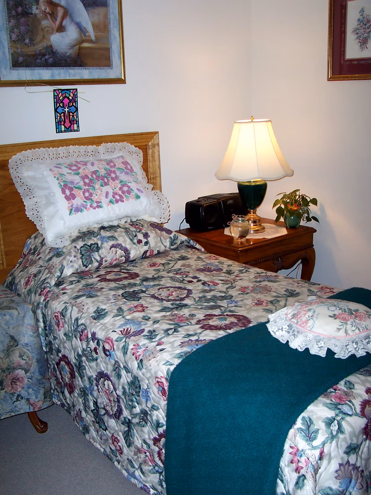 A cozy bedroom with a single bed covered in floral bedding and a teal blanket. There is a decorative pillow with lace on the bed. Next to the bed is a wooden nightstand with a green lamp, a small plant, a glass candle holder, and a black radio. The walls have framed artwork, including a painting of an angel and a floral print.