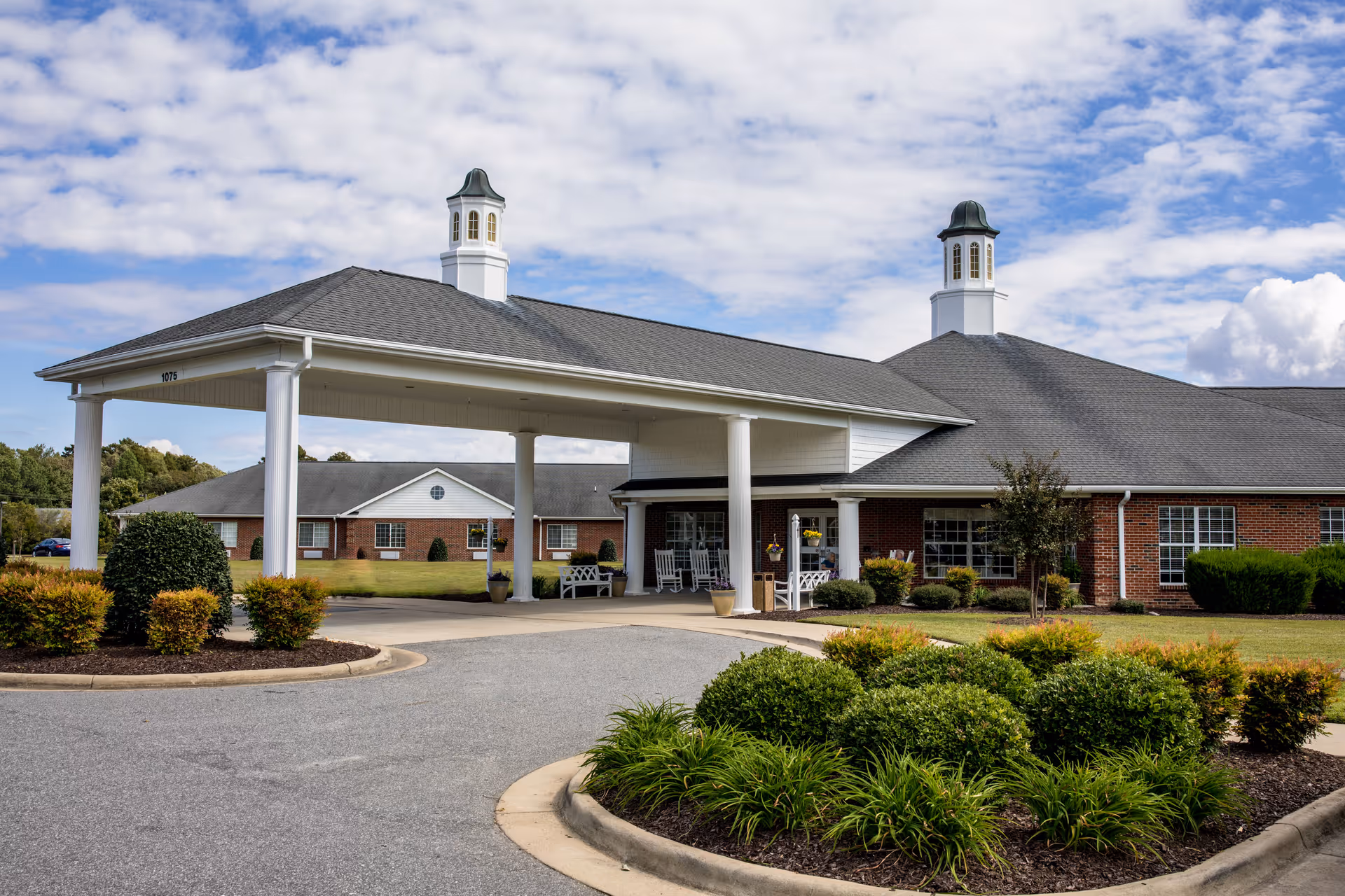 Exterior view of a single-story brick building with a large covered entrance supported by white columns. The building has two cupolas on the roof and is surrounded by well-maintained landscaping with bushes and grass under a partly cloudy sky.