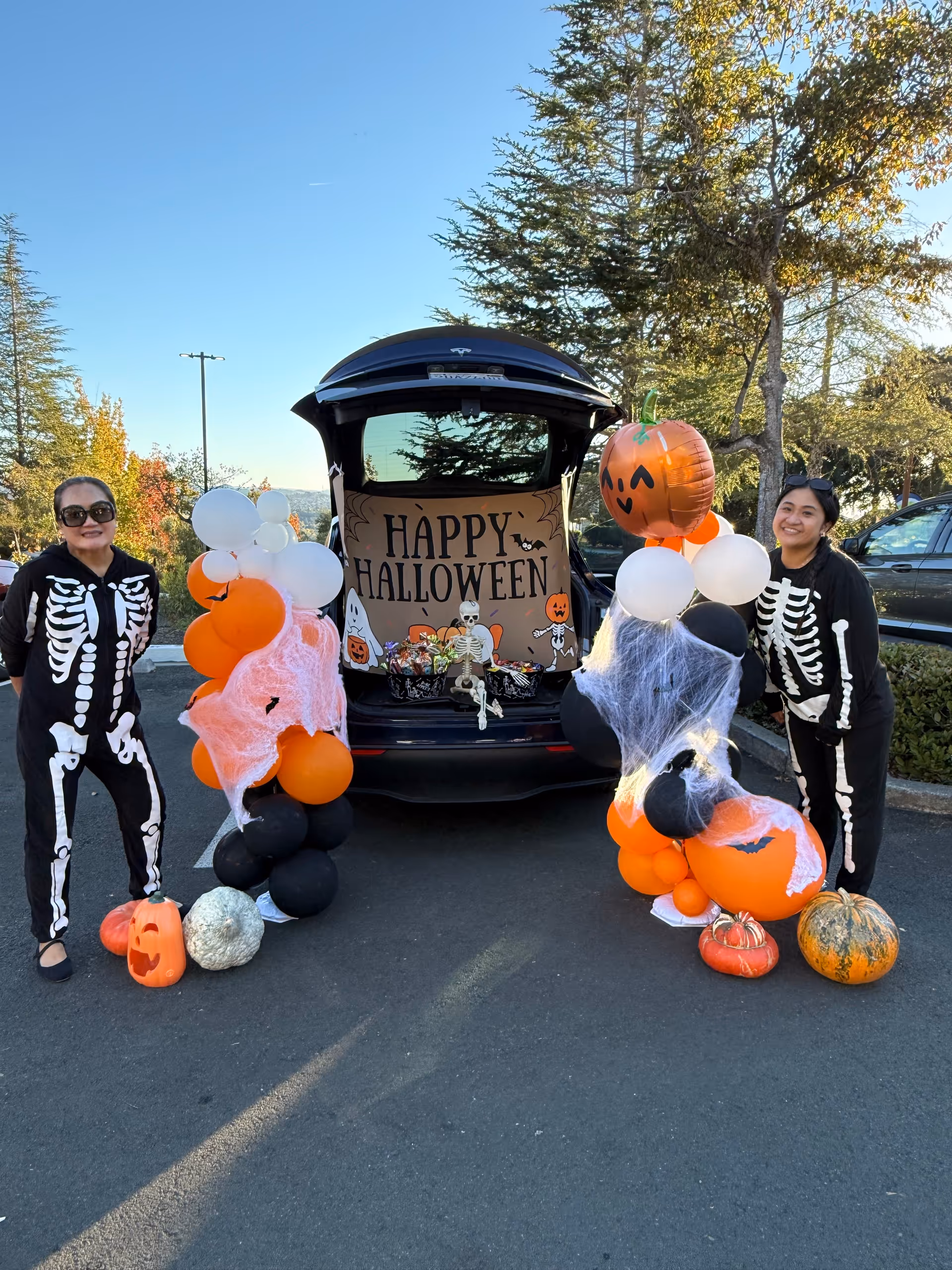 Two people in skeleton costumes pose beside a decorated Halloween trunk outdoors.