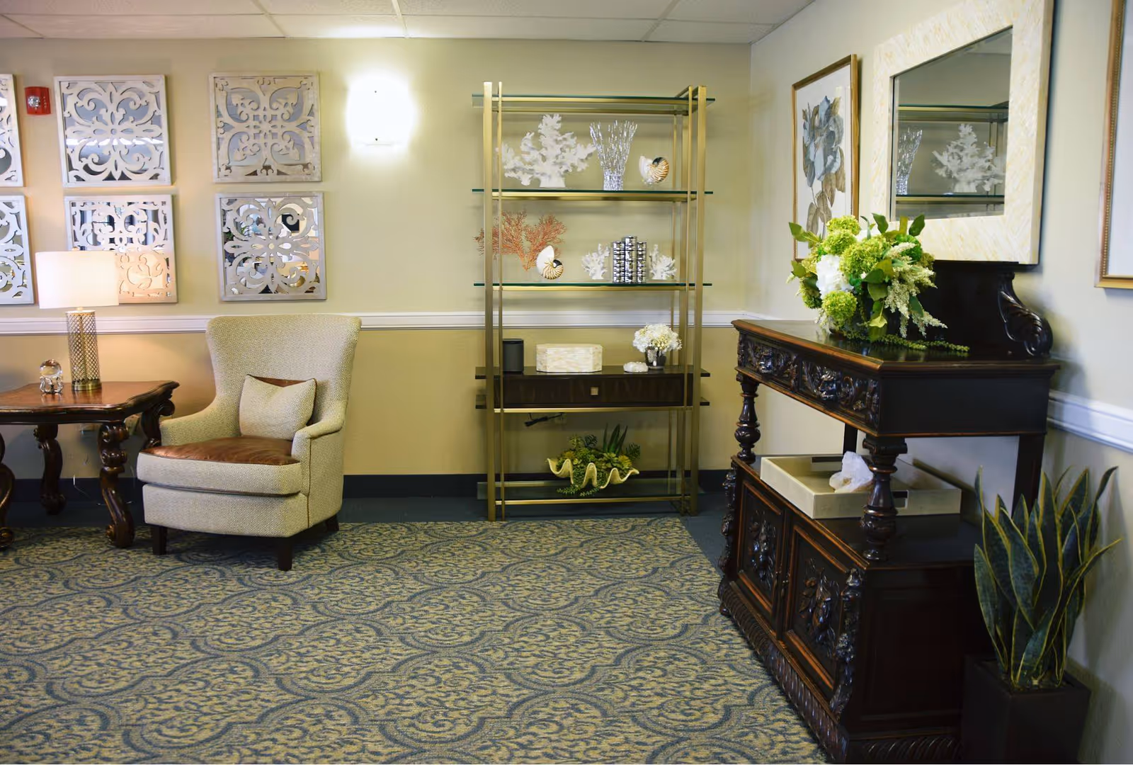 A cozy and elegantly decorated living room area in a senior living facility featuring a beige armchair with a brown cushion, a wooden side table with a lamp, a decorative shelving unit with glass shelves displaying coral and shell ornaments, a dark wooden console table with intricate carvings topped with a floral arrangement, a large mirror above the console, and a potted plant on the floor. The walls are light-colored with decorative wall art and the floor is covered with a patterned carpet.