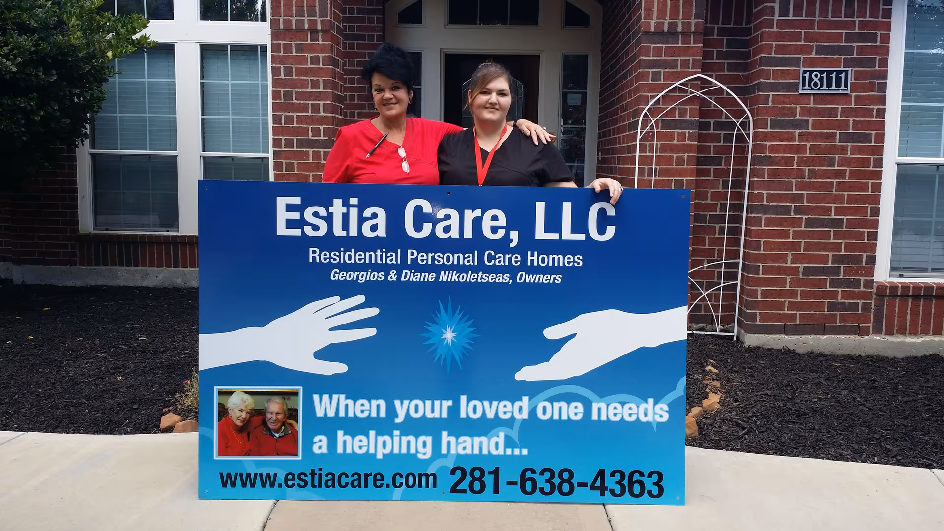 Two women stand behind a blue Estia Care, LLC sign in front of a brick building.
