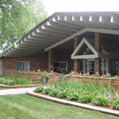Exterior view of a brick building with a large overhanging roof and a covered entrance. The entrance features a triangular architectural detail above the doorway. There are flower beds with green plants and flowers in front of the building, along with a well-maintained lawn and trees in the background.