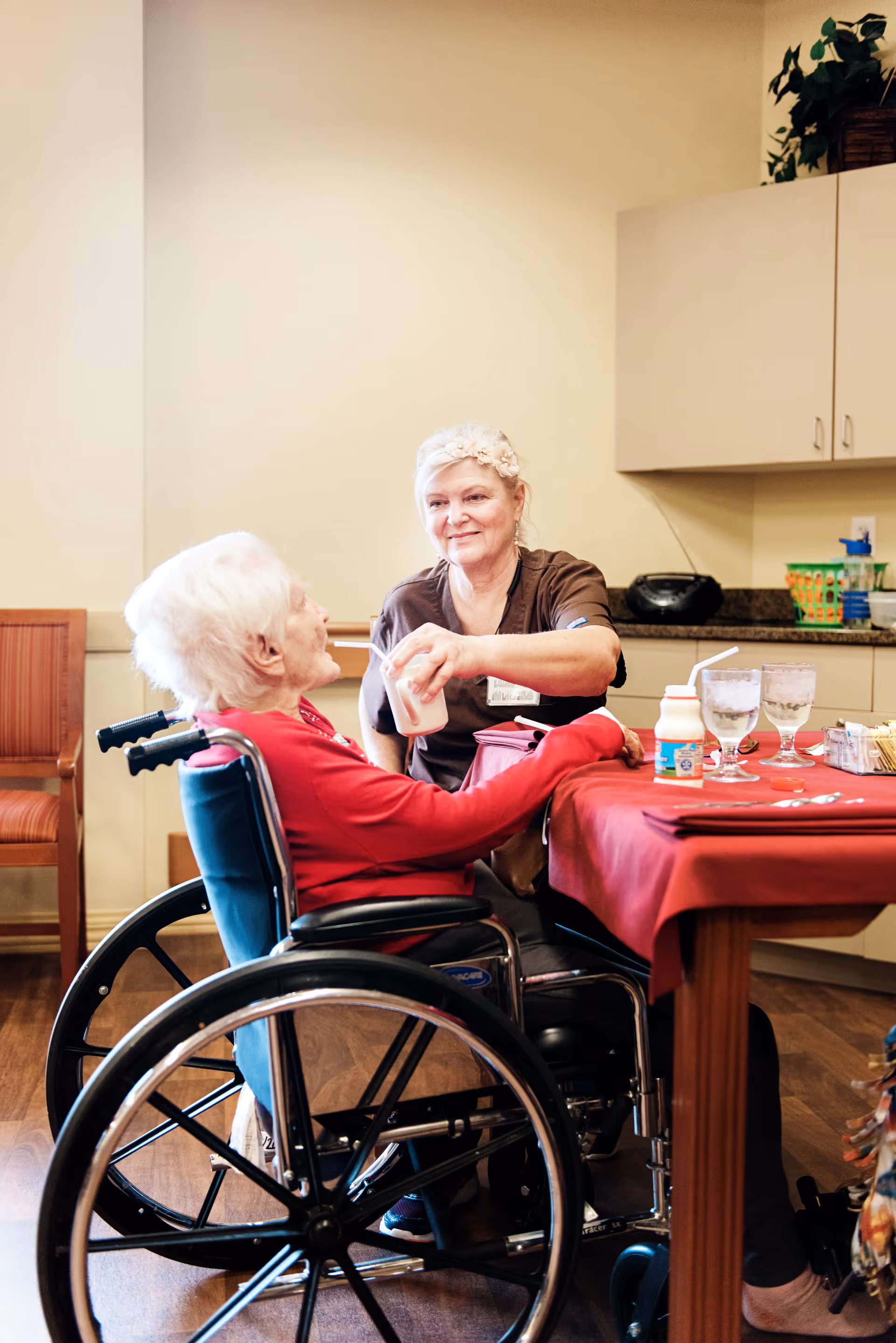 A caregiver is assisting an elderly woman in a wheelchair by helping her drink from a cup in a room with a table covered with a red tablecloth and kitchen cabinets in the background.