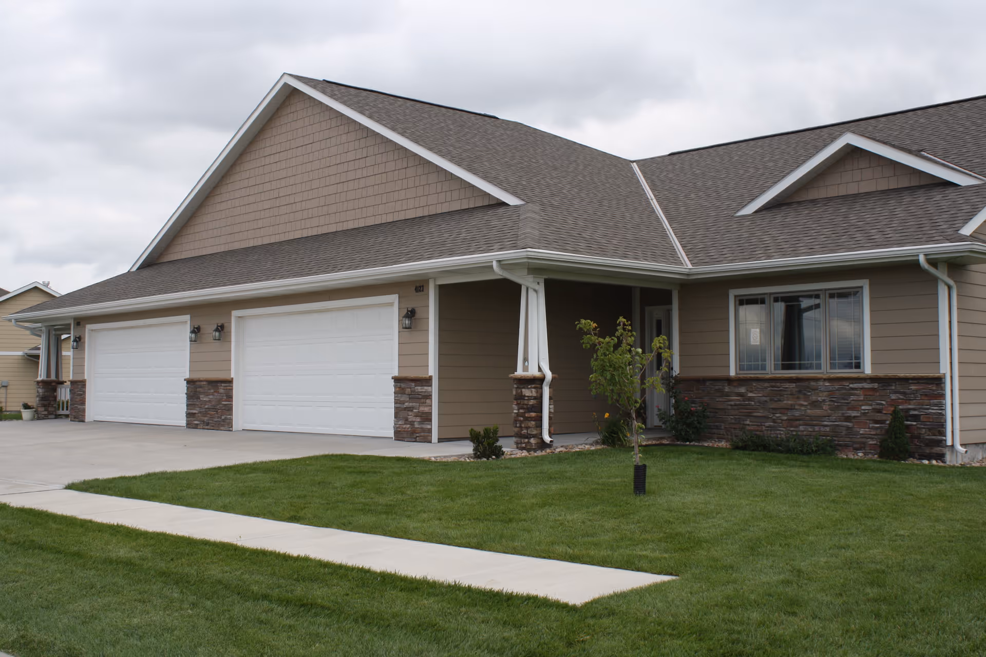 Exterior view of a single-story residential building with beige siding, stone accents, and a gray shingled roof. The building features two white garage doors, a small covered porch area, a young tree planted in the front lawn, and a concrete driveway and walkway.