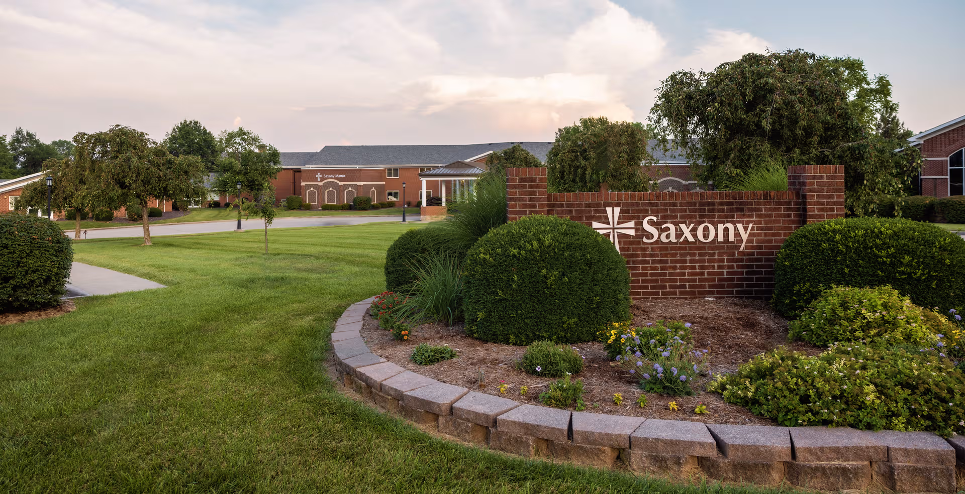 Brick Saxony sign and landscaped front lawn with pathway and trees at a senior living campus