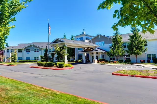 Exterior view of Clearwater Springs by Cogir, showing a large two-story building with a covered entrance, surrounded by trees and landscaping under a clear blue sky.