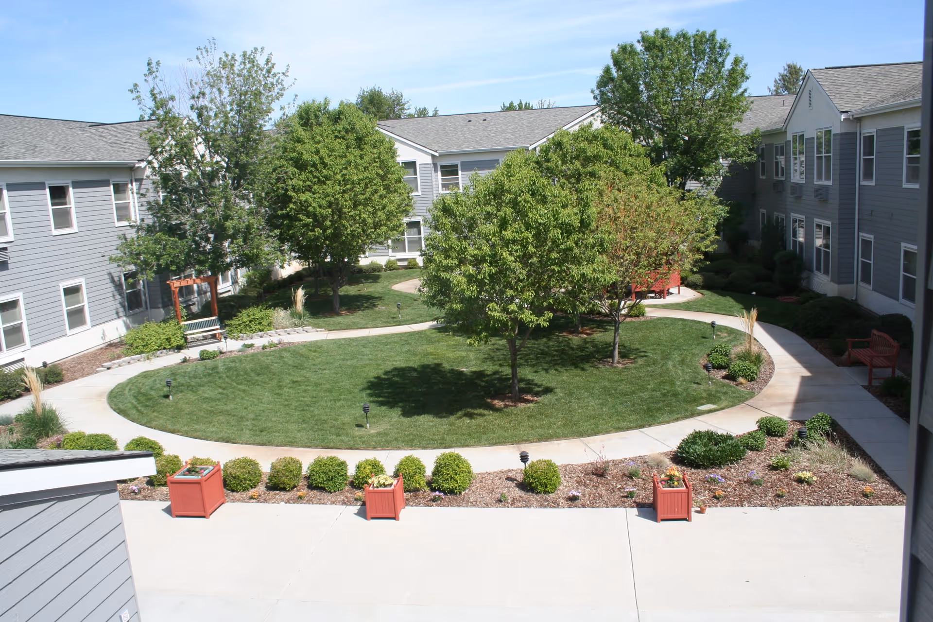 A landscaped courtyard area within a senior living facility featuring a circular grassy lawn surrounded by a concrete walkway. The courtyard is bordered by two-story gray buildings with white trim. There are several trees, shrubs, and red planters with flowers, along with benches placed along the walkway.
