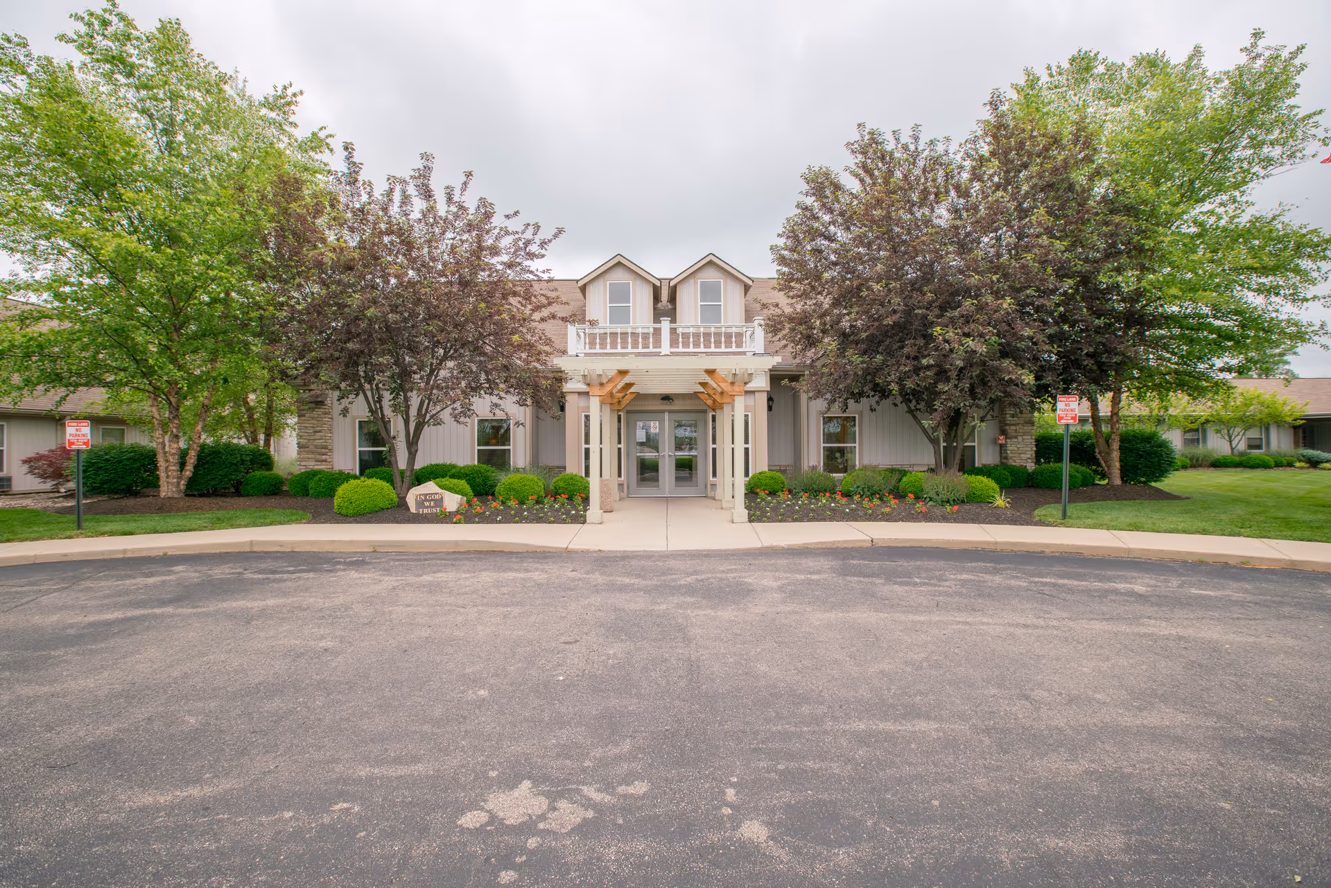 Front exterior view of a single-story building with a covered entrance, surrounded by well-maintained landscaping including trees, bushes, and flowers under a cloudy sky.