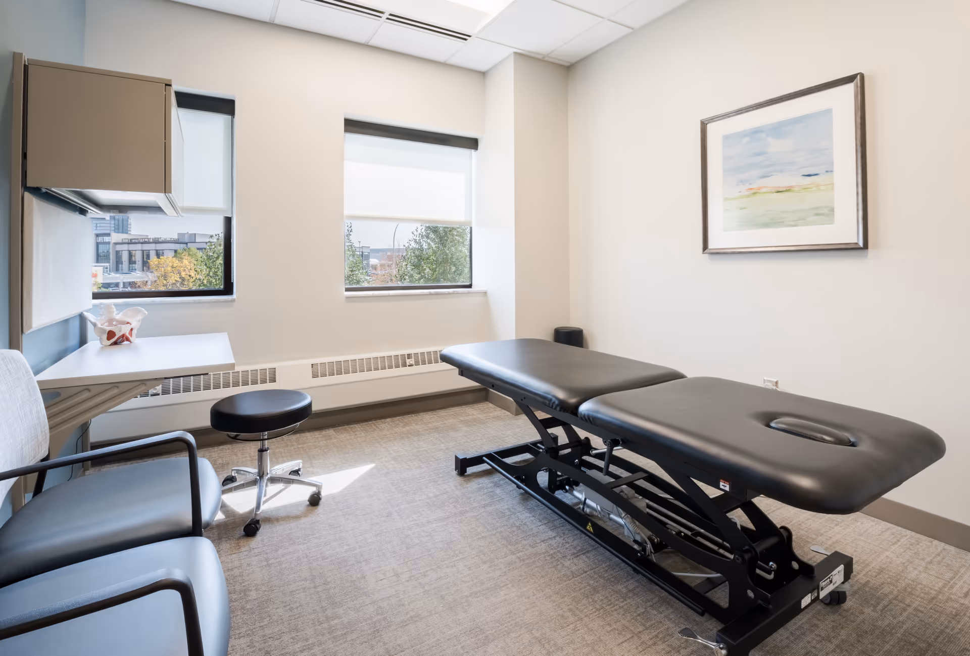 A clinical treatment room with an adjustable therapy table, stools, chairs, and large windows.