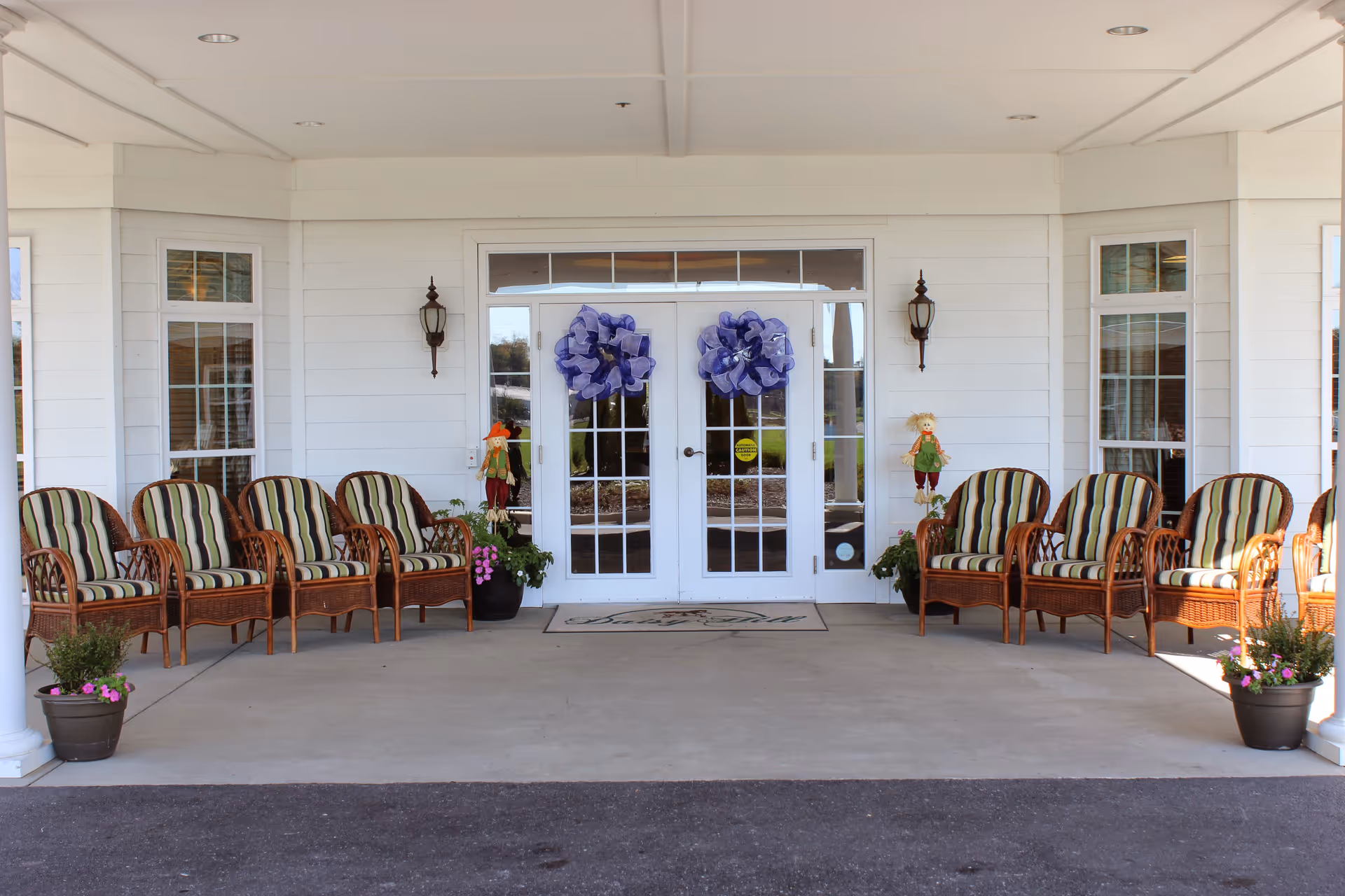 Entrance area of a senior living facility with white double doors decorated with large purple bows. There are six wicker chairs with striped cushions arranged on each side of the entrance, along with potted plants and small scarecrow decorations. The building exterior is white with windows on either side of the doors.