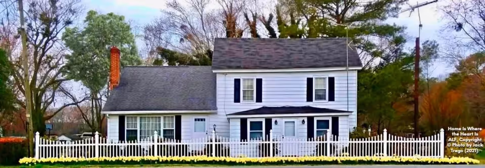 A two-story white house with black shutters and a gray roof, surrounded by a white picket fence and yellow flowers in front. Trees with autumn foliage are visible in the background under a partly cloudy sky.