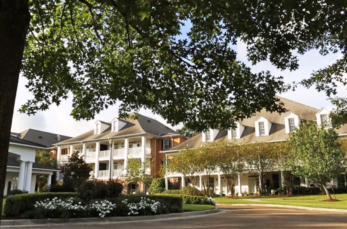 Front view of a multi-story senior living building with balconies, landscaped grounds, and large trees.