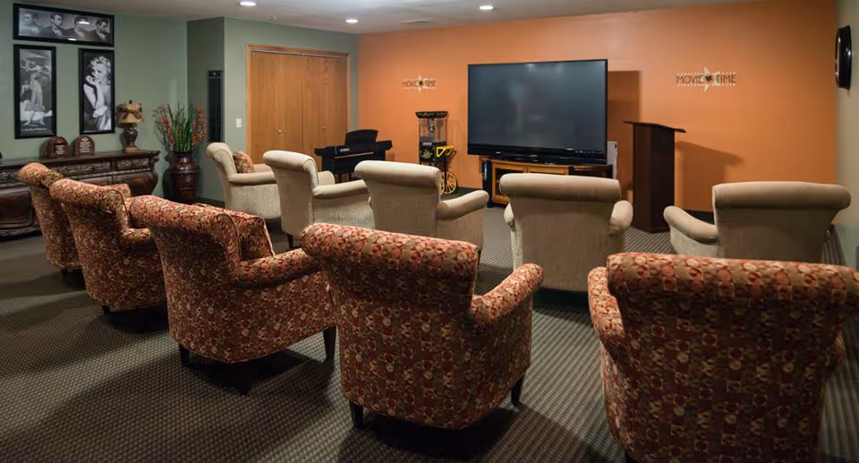 A cozy supportive living facility room set up as a small movie theater with two rows of cushioned armchairs facing a large flat-screen TV mounted on an orange wall. The room has a carpeted floor, decorative framed photos on one wall, a popcorn machine, and a small podium.