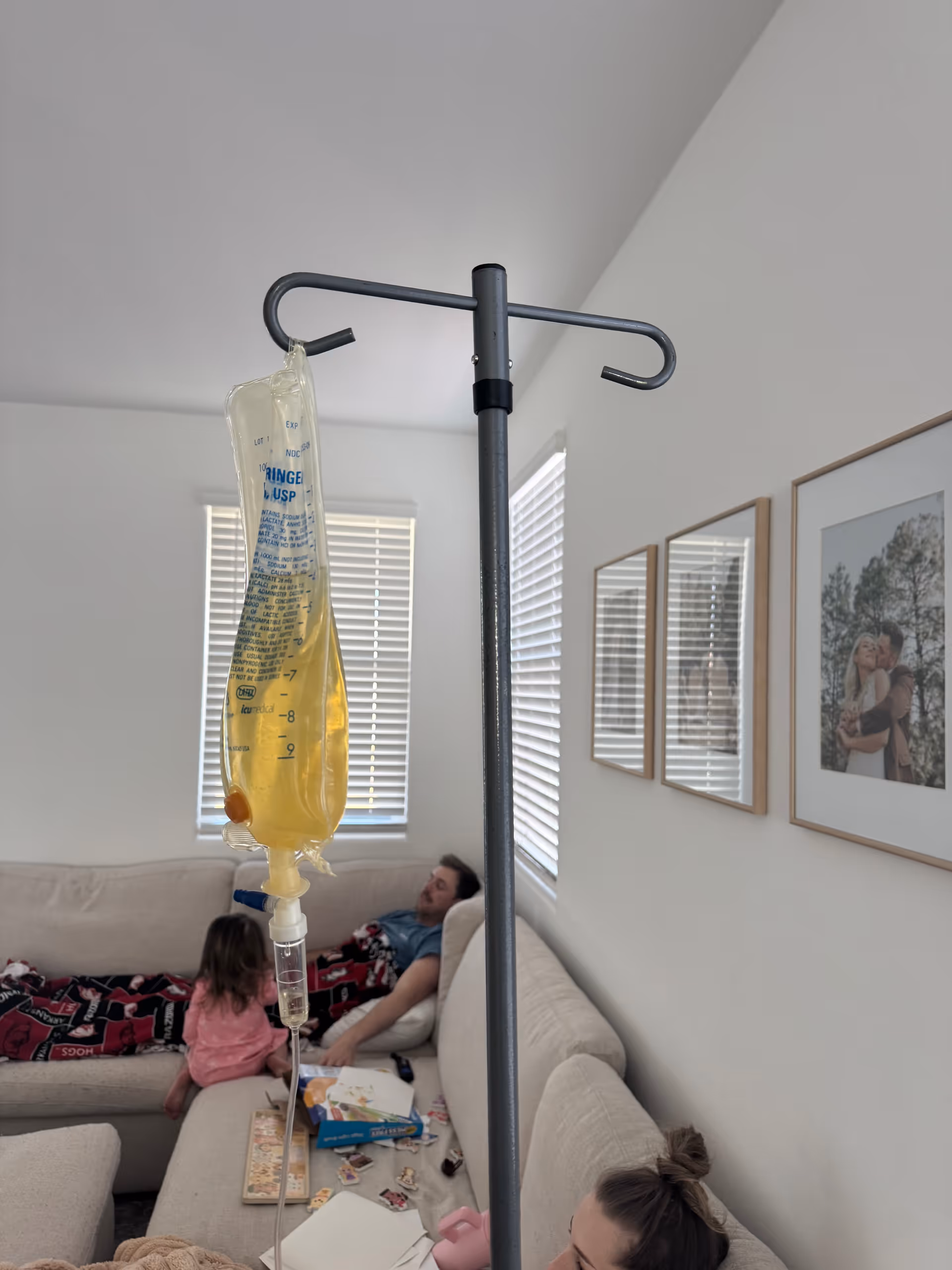IV drip bag on a stand in a bright living room, with people on a couch.