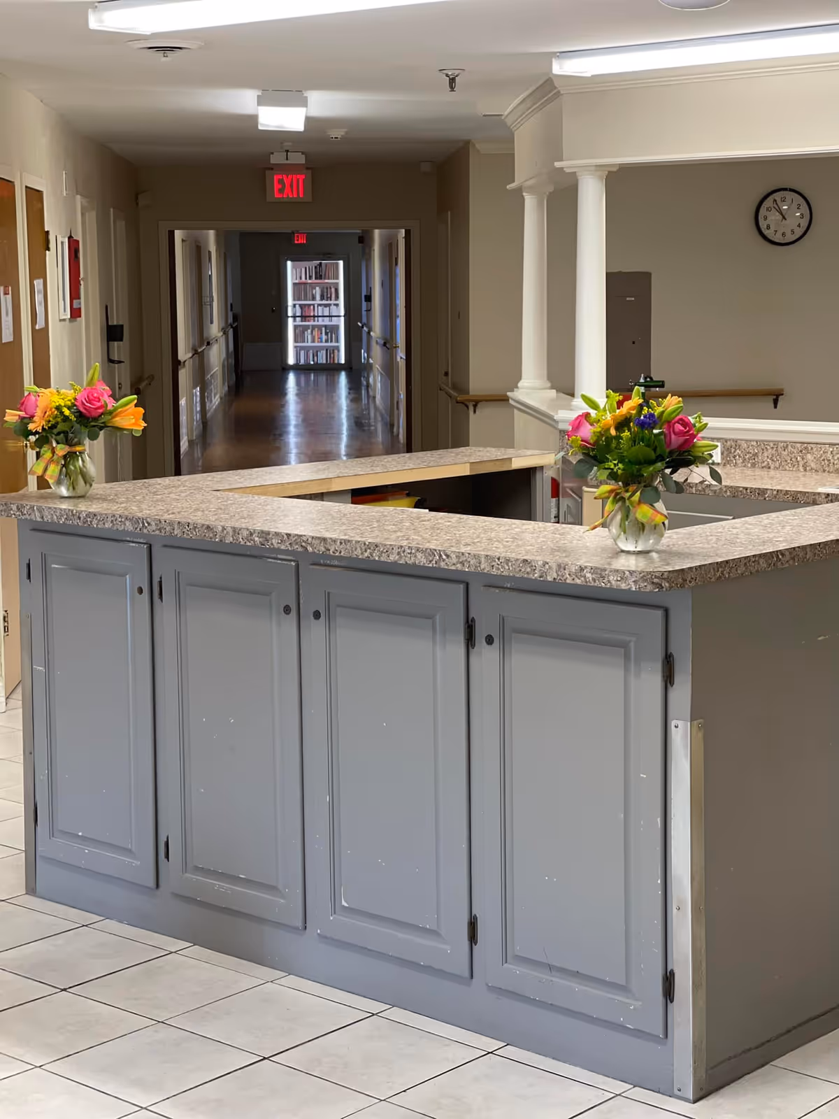 Interior reception desk with gray cabinets, flowers, clock, and long hallway in background.