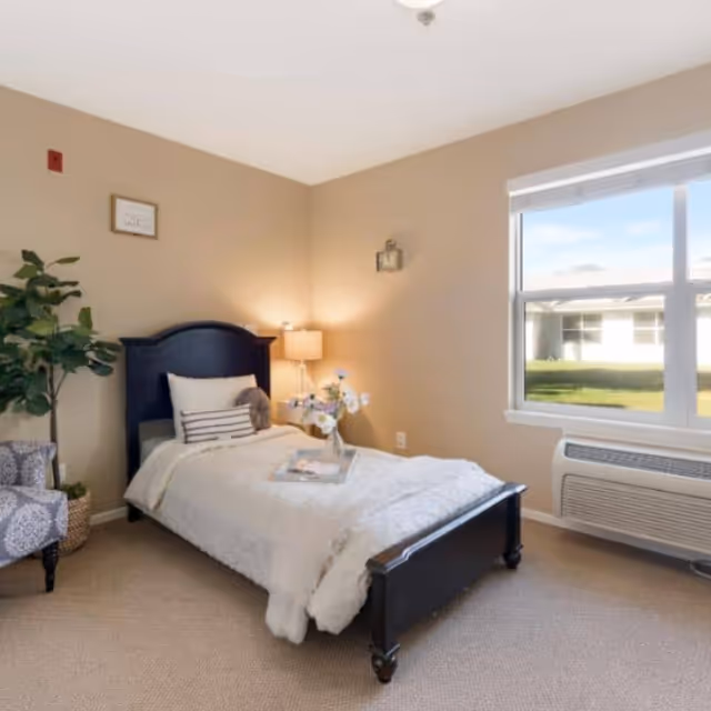 Cozy bedroom in a senior living facility featuring a dark wood bed, window, and plants.