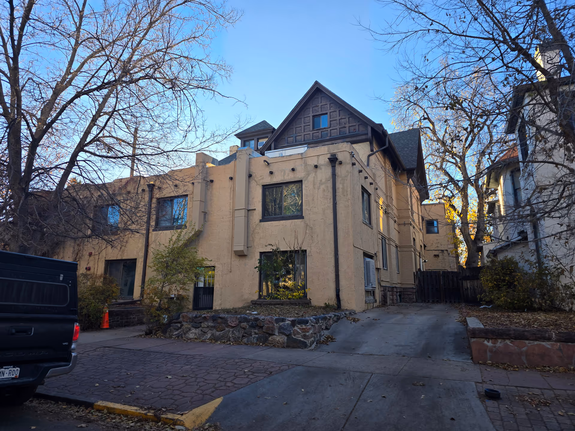 Exterior view of a multi-story beige building with a steep roof and several windows, surrounded by leafless trees and a driveway with a black pickup truck parked on the left side.