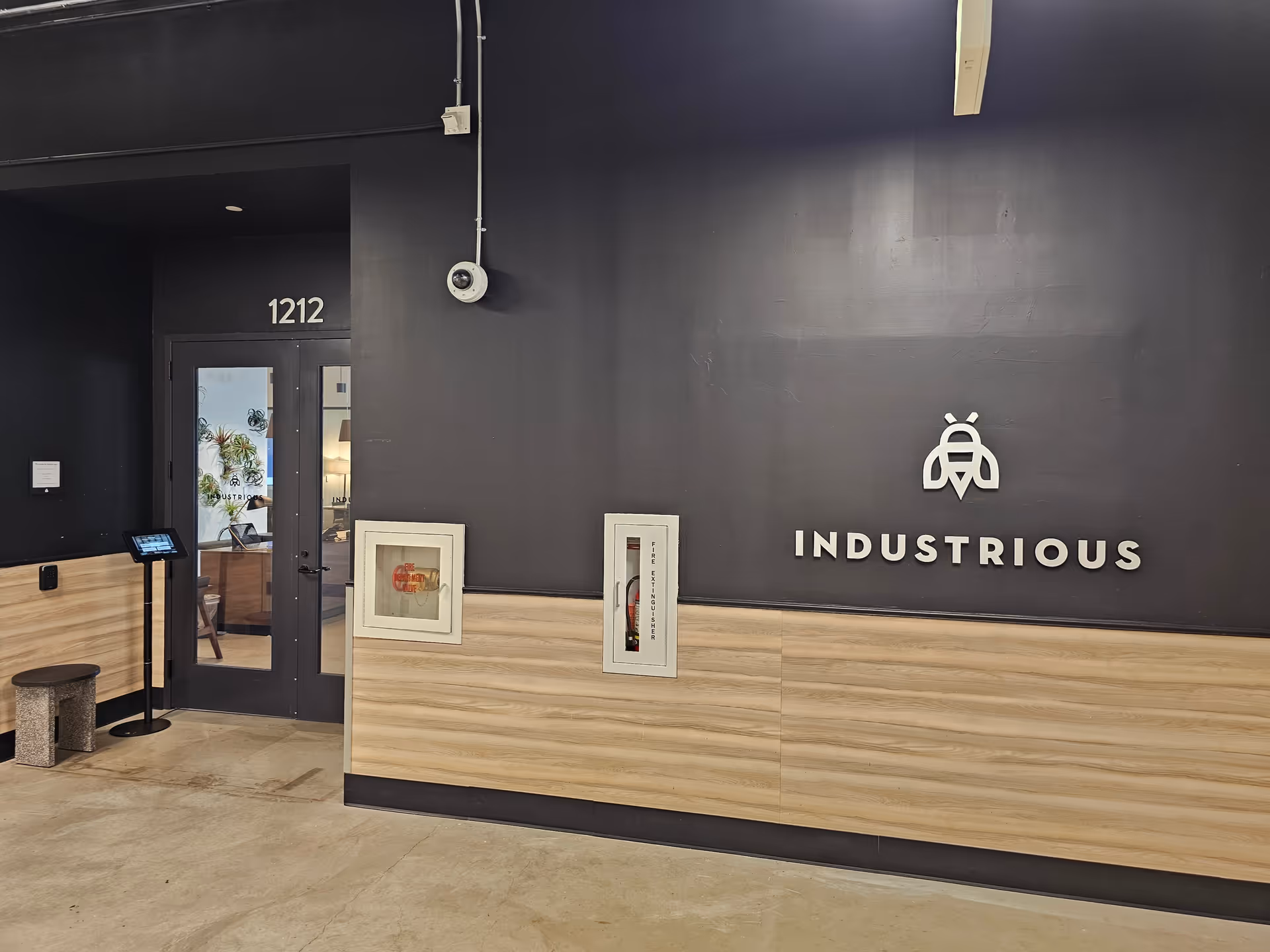 Dark interior hallway with INDUSTRIOUS logo, wooden paneling, and a glass door labeled 1212.