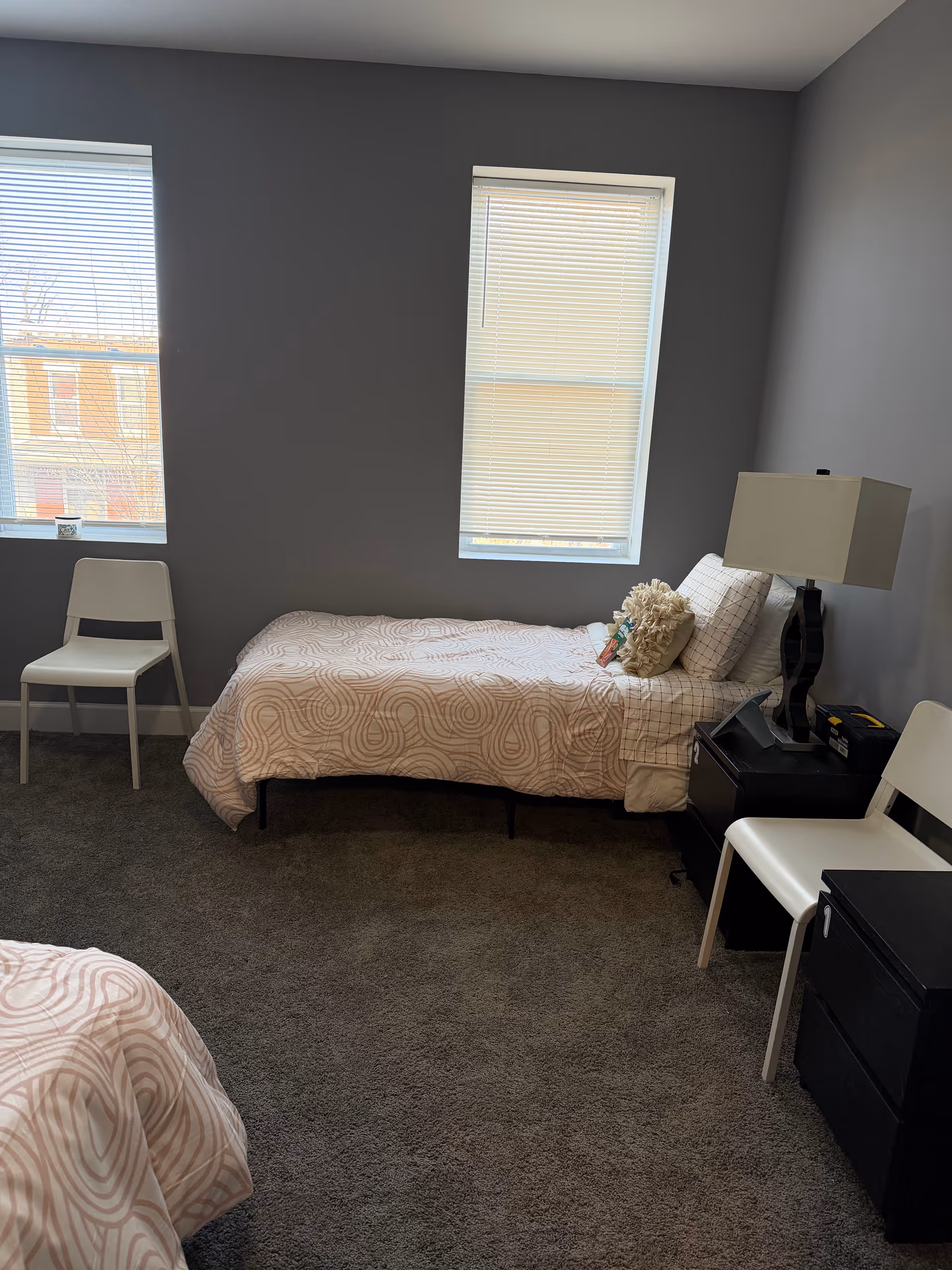 A gray-walled bedroom with two twin beds, two windows, chairs, and a nightstand.