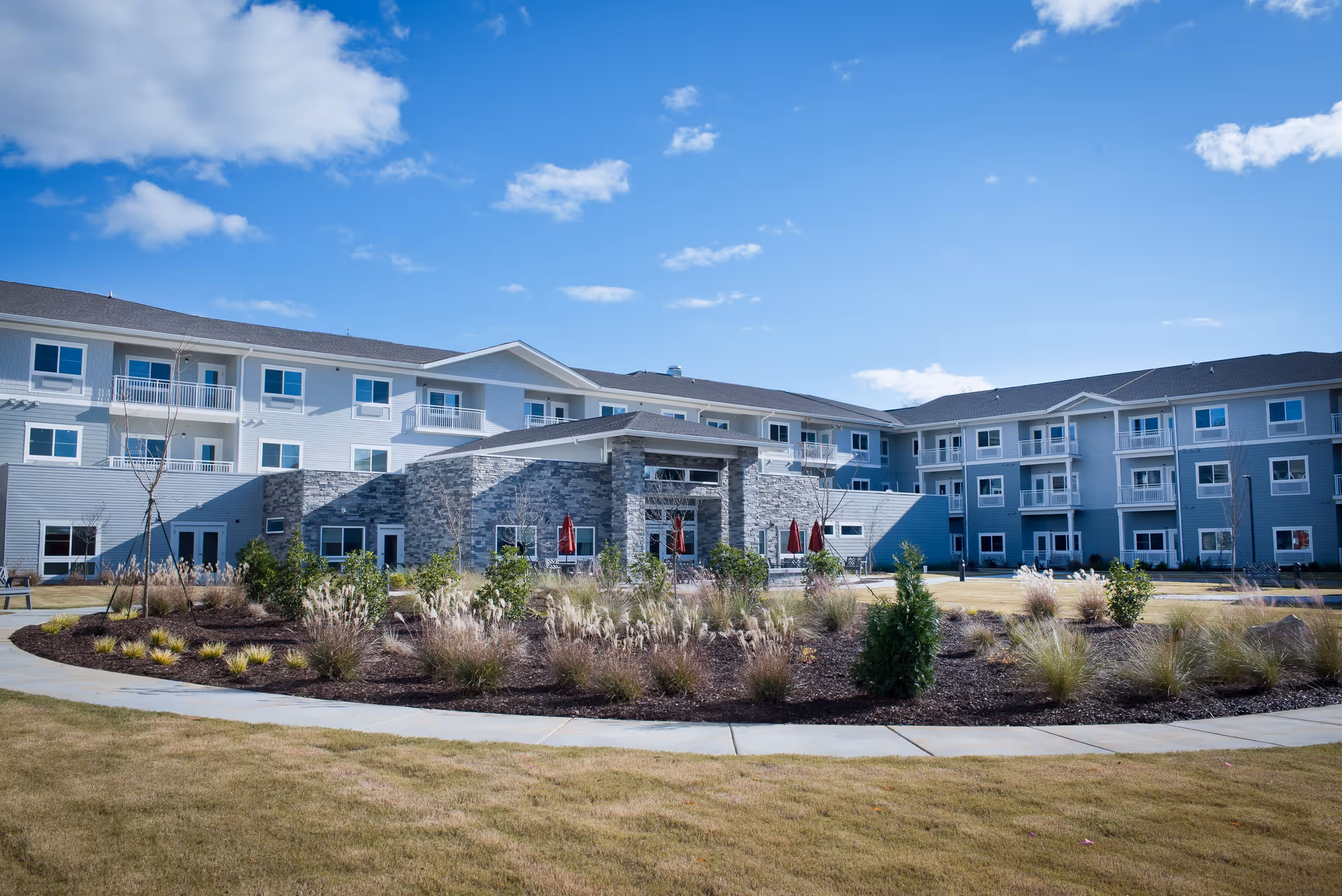 Exterior view of a modern three-story retirement community building with gray siding and stone accents, surrounded by landscaped gardens and a curved sidewalk under a blue sky with scattered clouds.