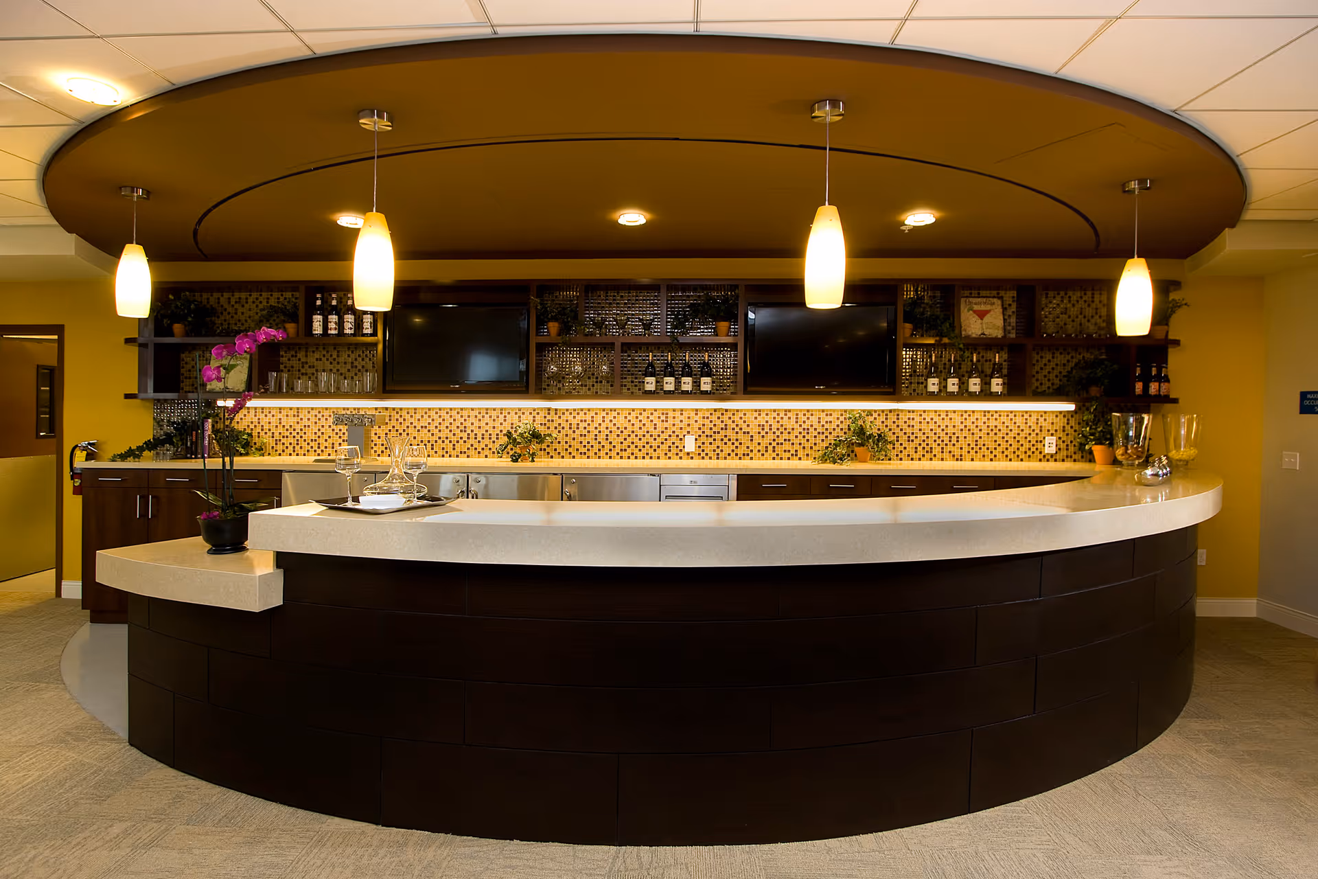 Curved dark-wood counter and white countertop with hanging pendant lights, mosaic tile backsplash, shelving and two wall TVs in a communal kitchen/lounge area.