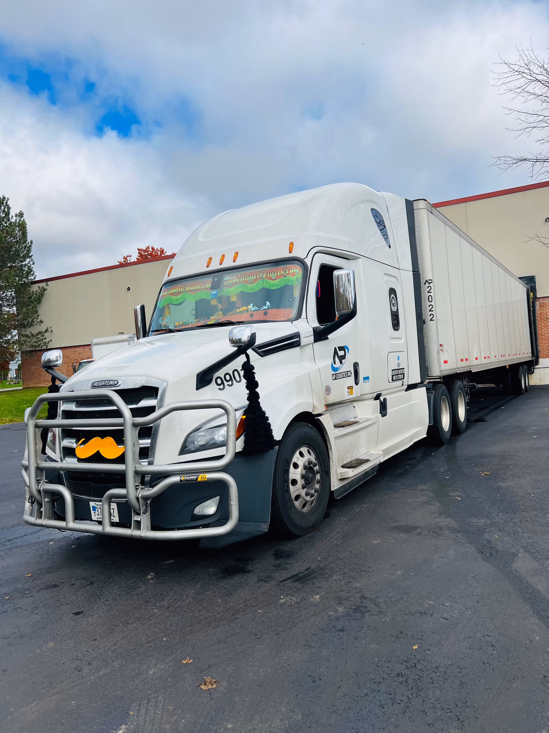 White semi-truck with long trailer parked in a lot in front of a beige building.