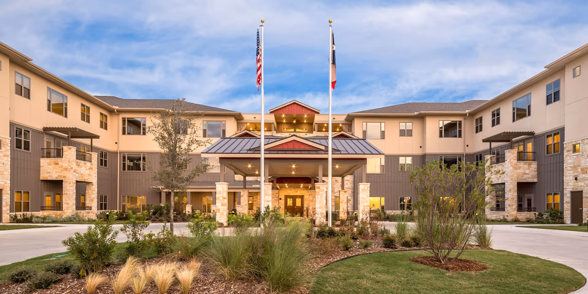 Front exterior view of Village on the Park Plano, a multi-story senior living facility with beige and gray walls, stone accents, and a covered entrance. Two flagpoles with the American and Texas flags stand in front of the building, surrounded by landscaped greenery and a circular driveway.