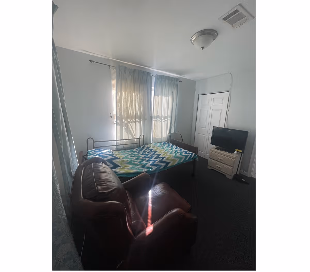 A bedroom in a care facility with bed, curtains, leather chair, dresser, and TV.