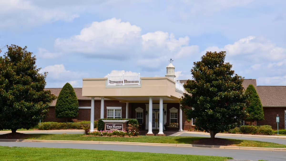 Front exterior view of The Haven & The Laurels in the Village at Carolina Place building with a covered entrance supported by white columns, surrounded by neatly trimmed trees and landscaping under a partly cloudy sky.